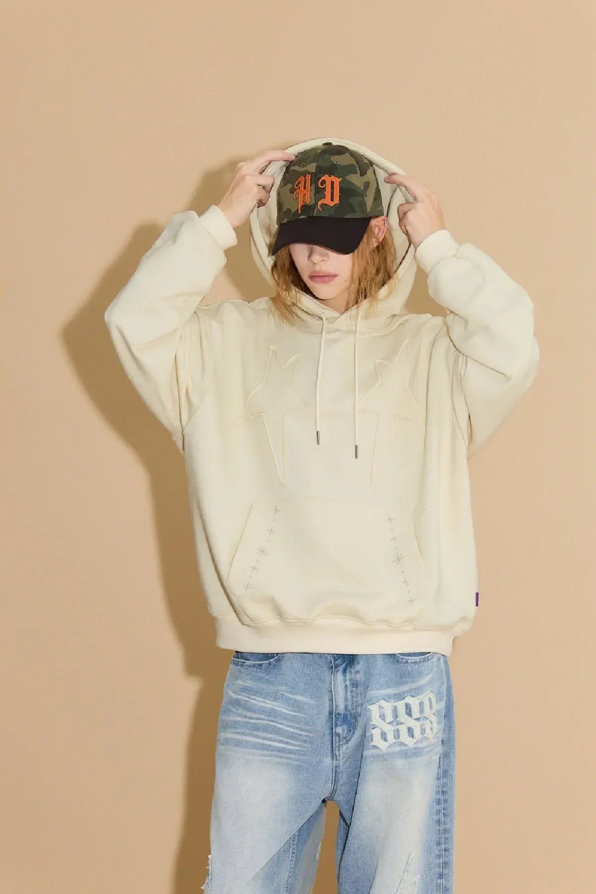 Person wearing ivory star applique hoodie with light blue jeans and camo baseball cap with orange logo