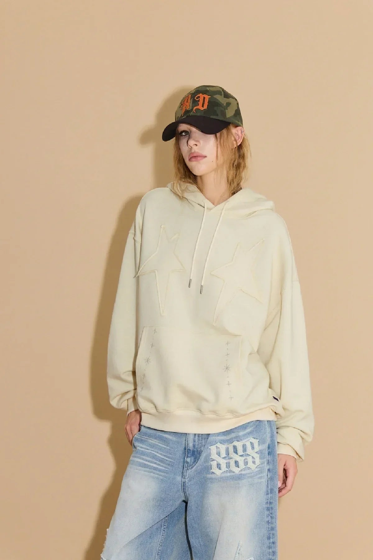 Model wearing ivory hoodie with star appliqué and blue jeans with white embroidery against beige background