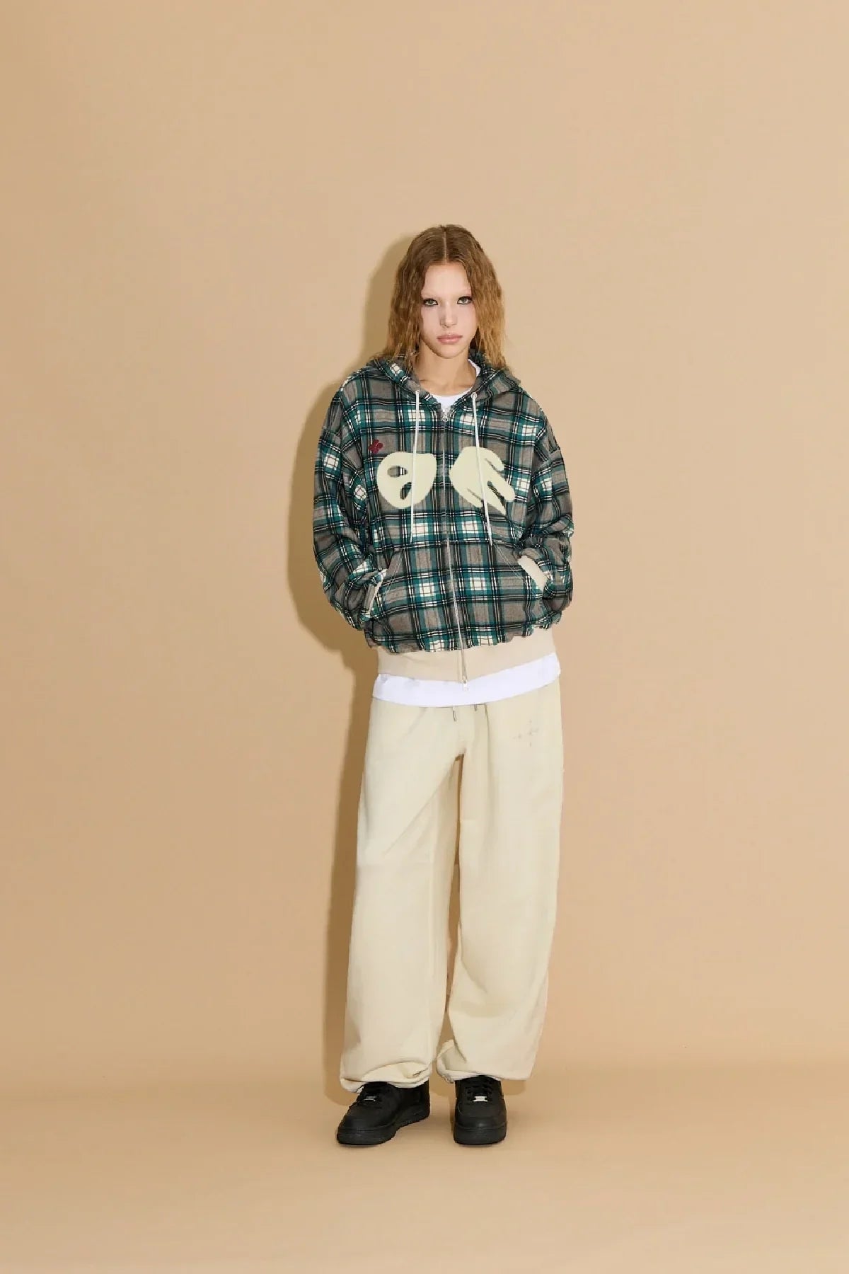 Model wearing green plaid hoodie with white hand appliques, cream wide-leg pants, and black shoes against beige background