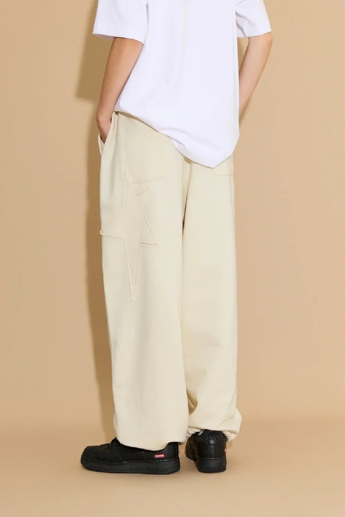 Model wearing ivory wide-leg sweatpants with star appliqué and black sneakers against beige background