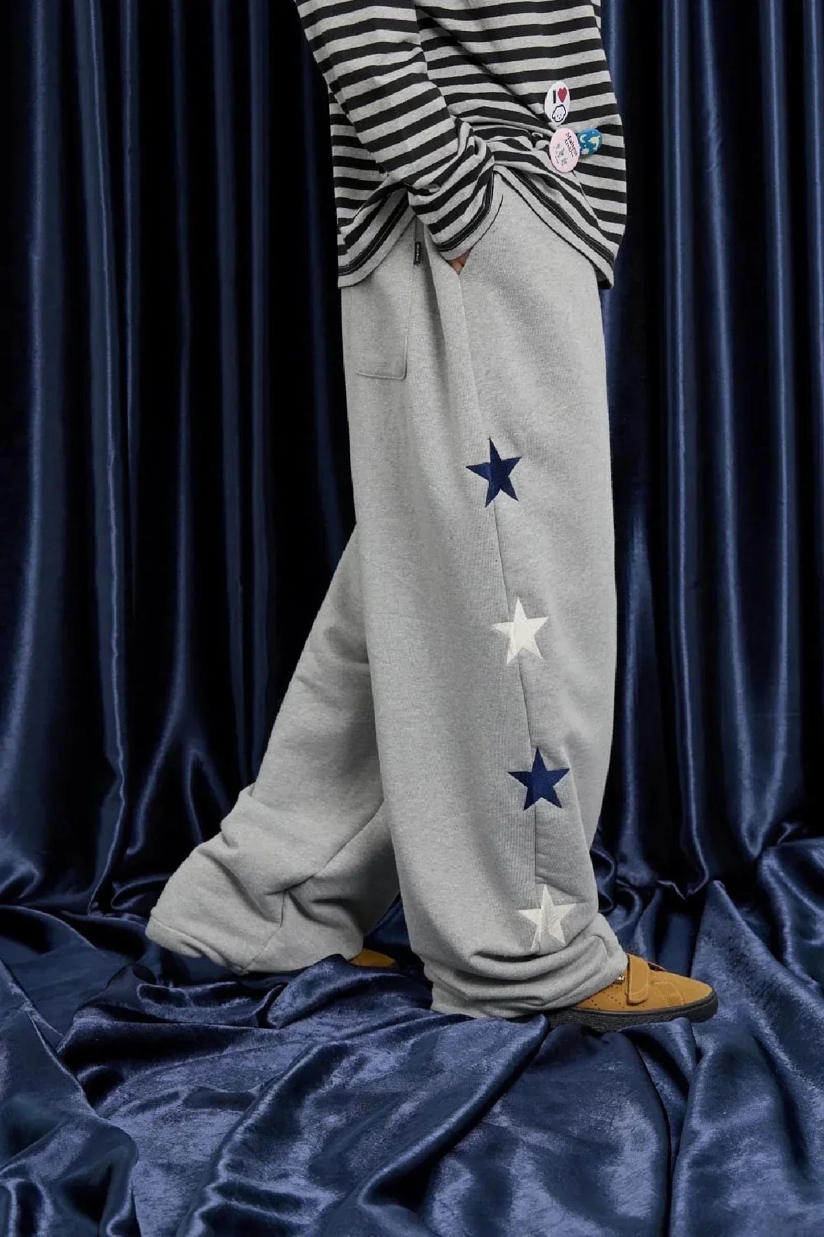 Person wearing loose grey sweatpants with navy and white star appliques on the side, paired with striped shirt and brown shoes on blue velvet