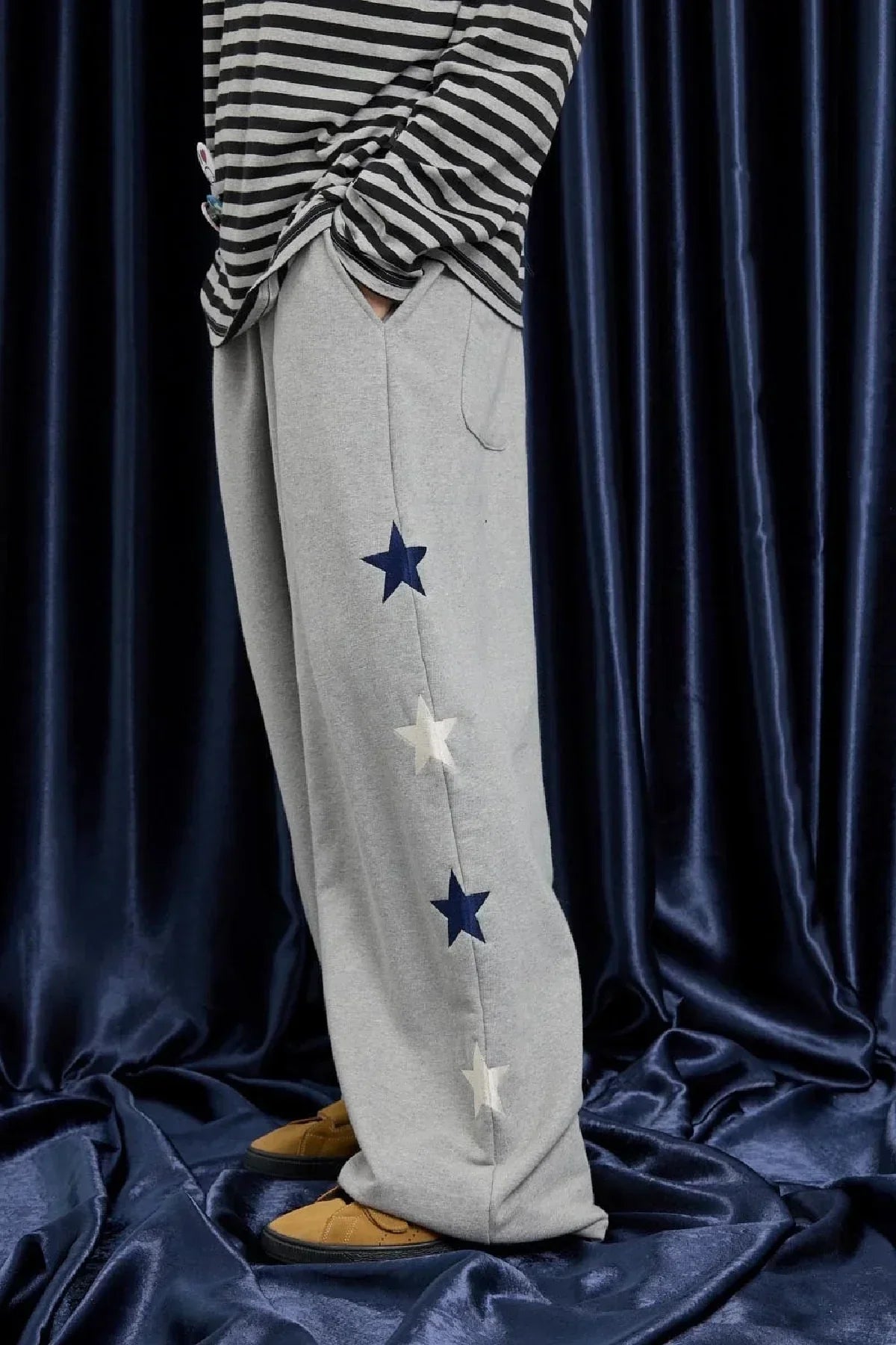 Side view of grey sweatpants with navy and white star patterns worn with striped shirt and mustard shoes on blue fabric background