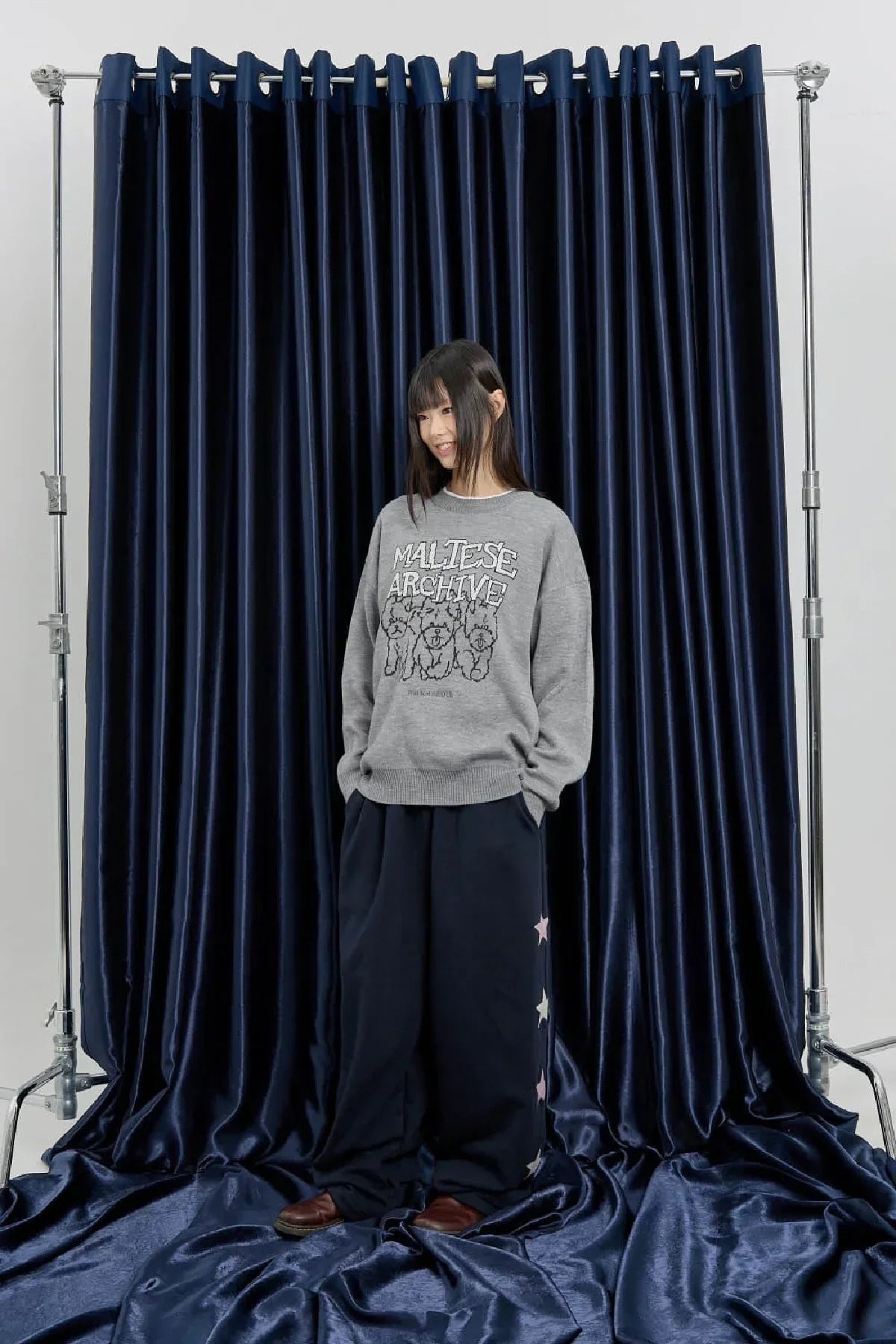 Woman wearing a gray graphic MALTESE ARCHIVE sweater and navy star-patterned wide pants on velvet backdrop