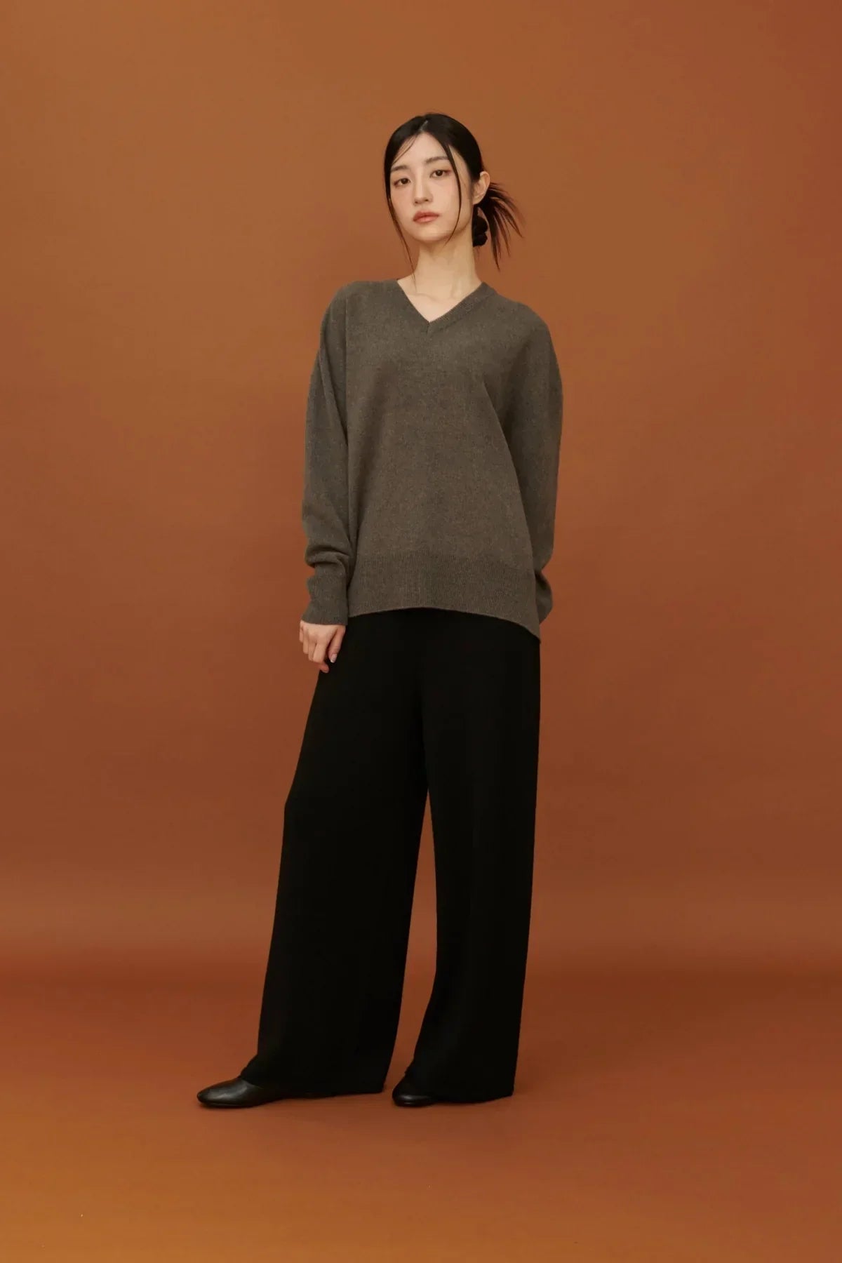 Woman in gray V-neck sweater and black wide-leg pants against brown backdrop