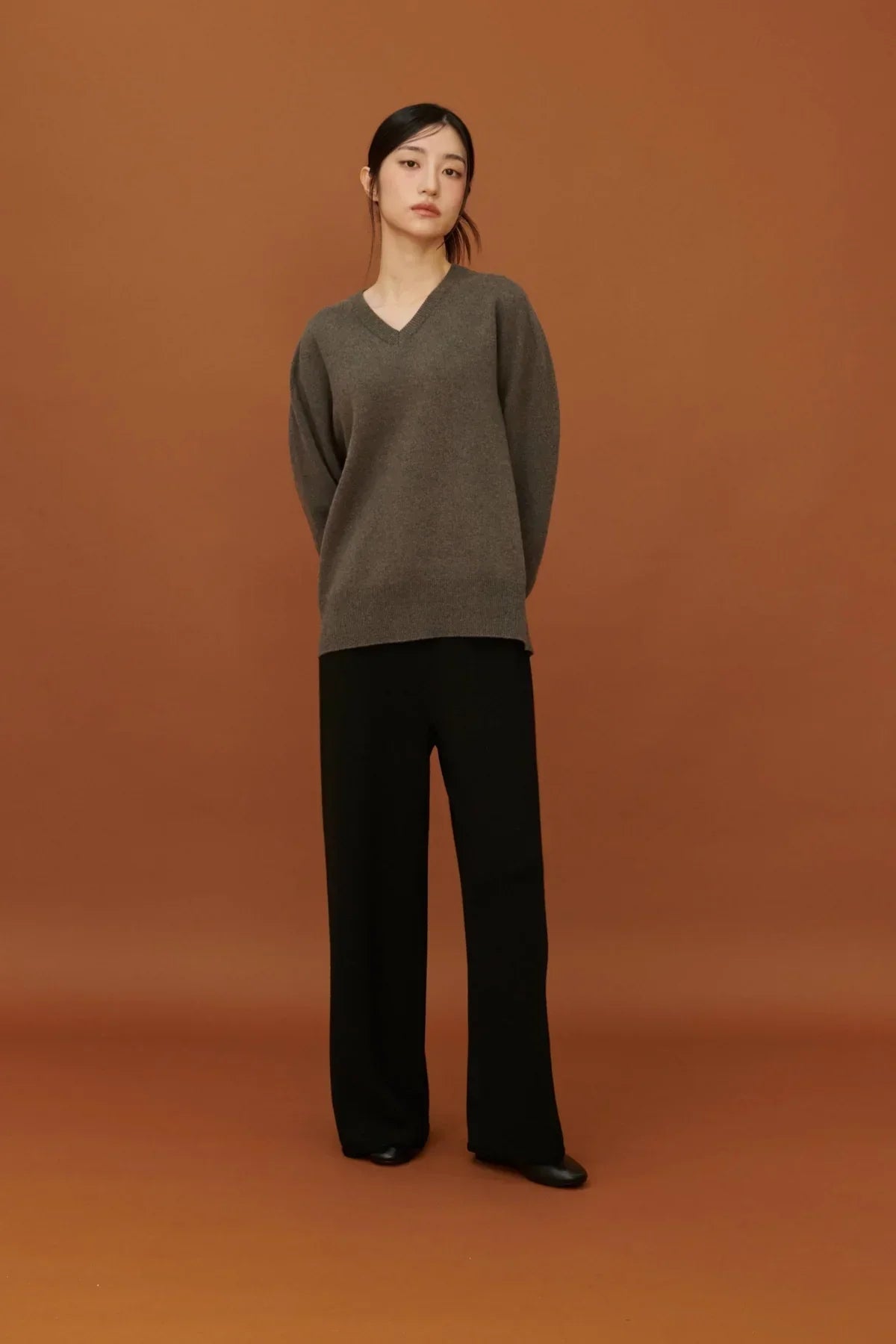 Woman wearing grey V-neck sweater and black straight fit knit pants on brown background