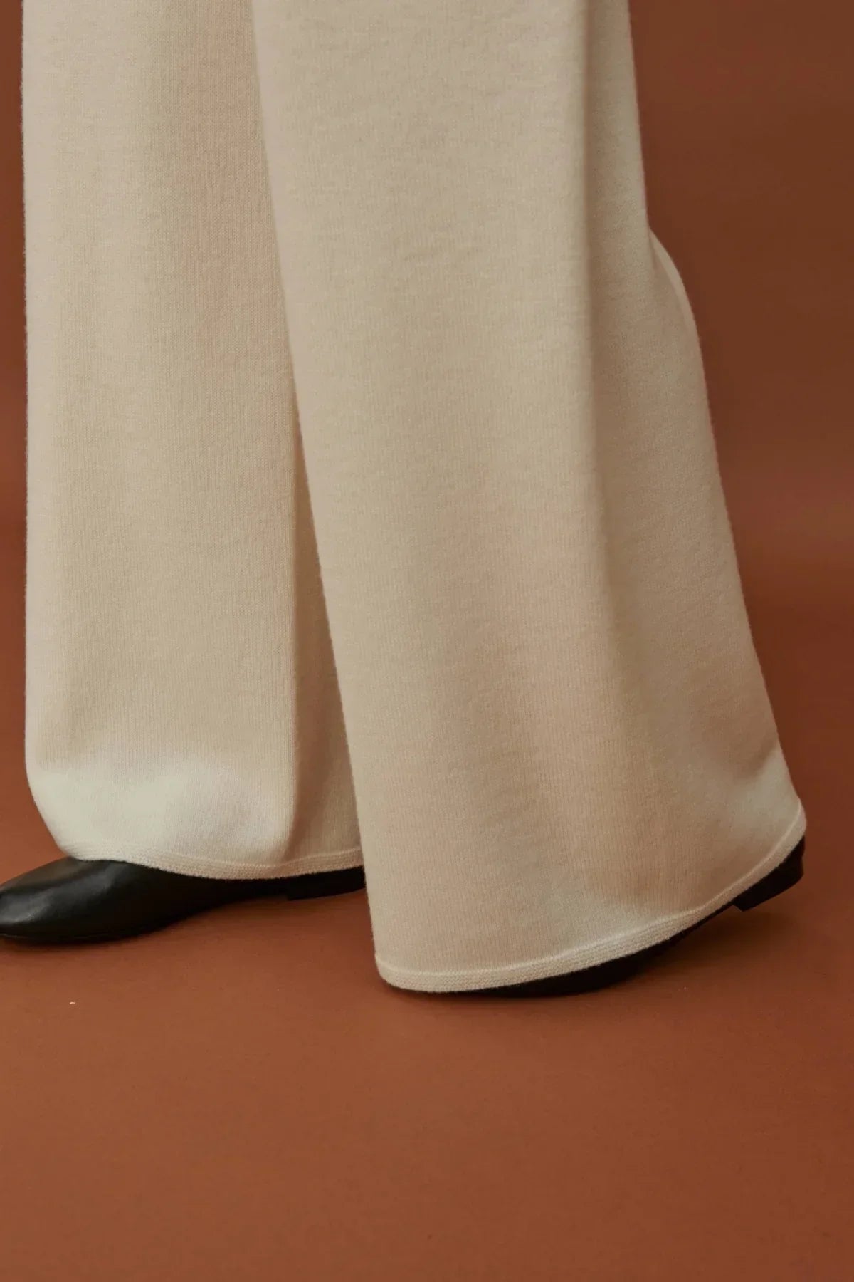 Close-up of white straight fit knit pants covering black shoes on brown floor