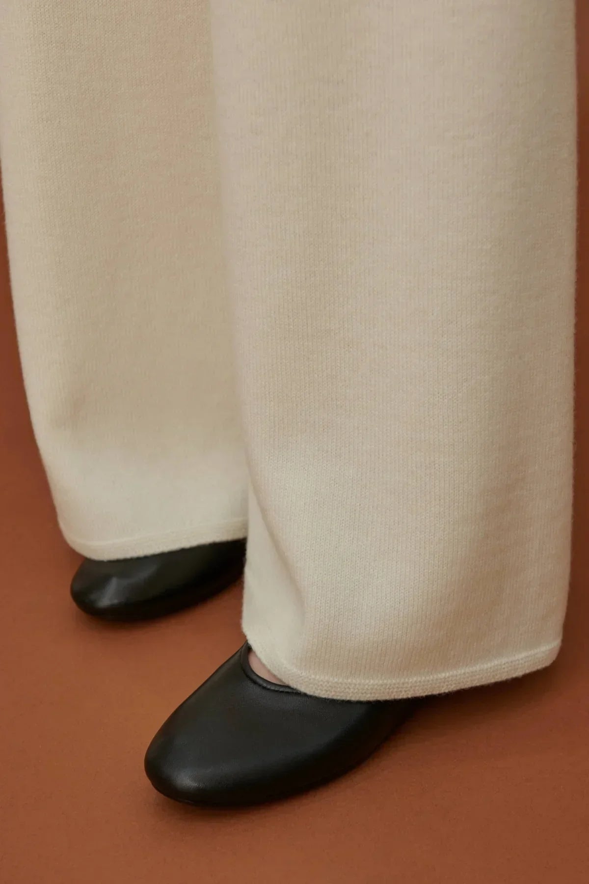 Close-up of white straight-fit knit pants paired with black slip-on shoes on brown floor