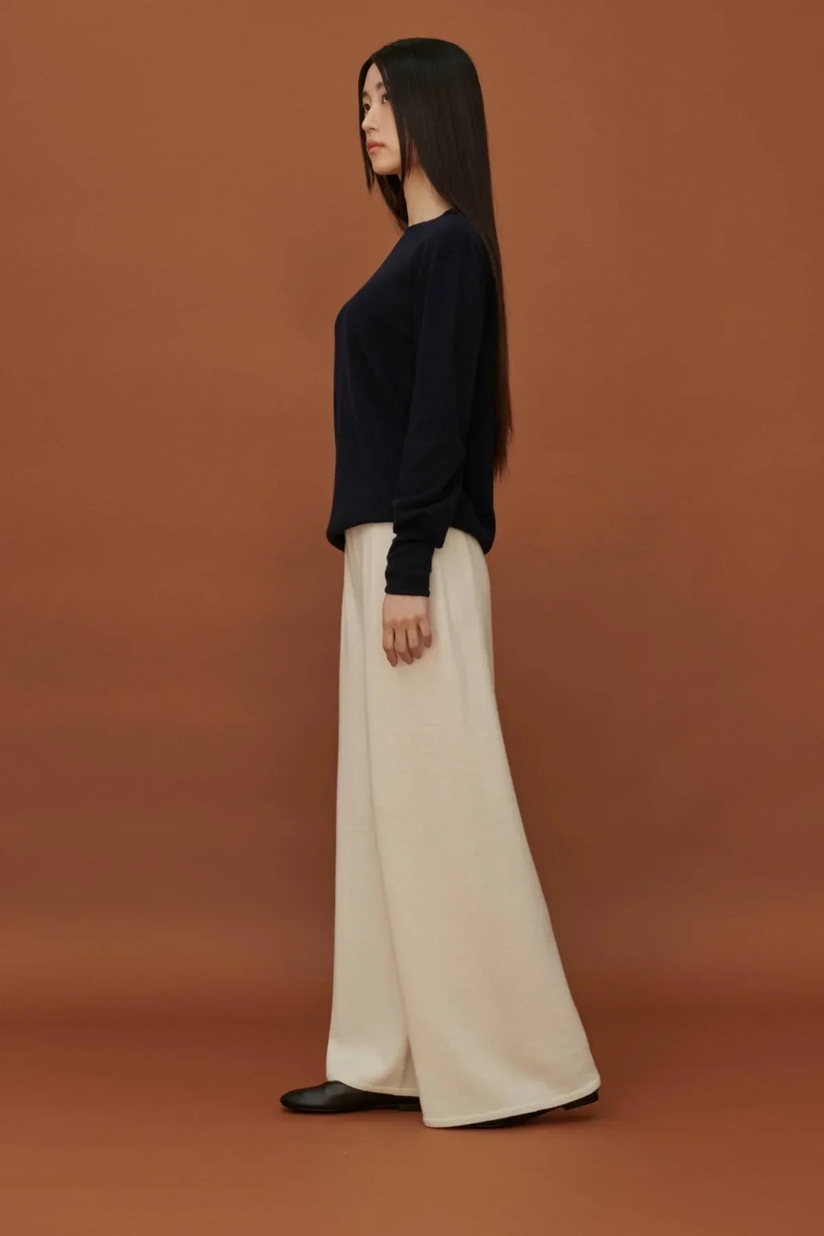 Side view of woman wearing black long-sleeve top and white wide-leg knit pants against brown background