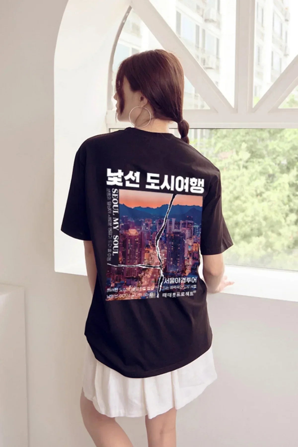 Woman wearing black Seoul cityscape graphic t-shirt with Korean text, standing by white window