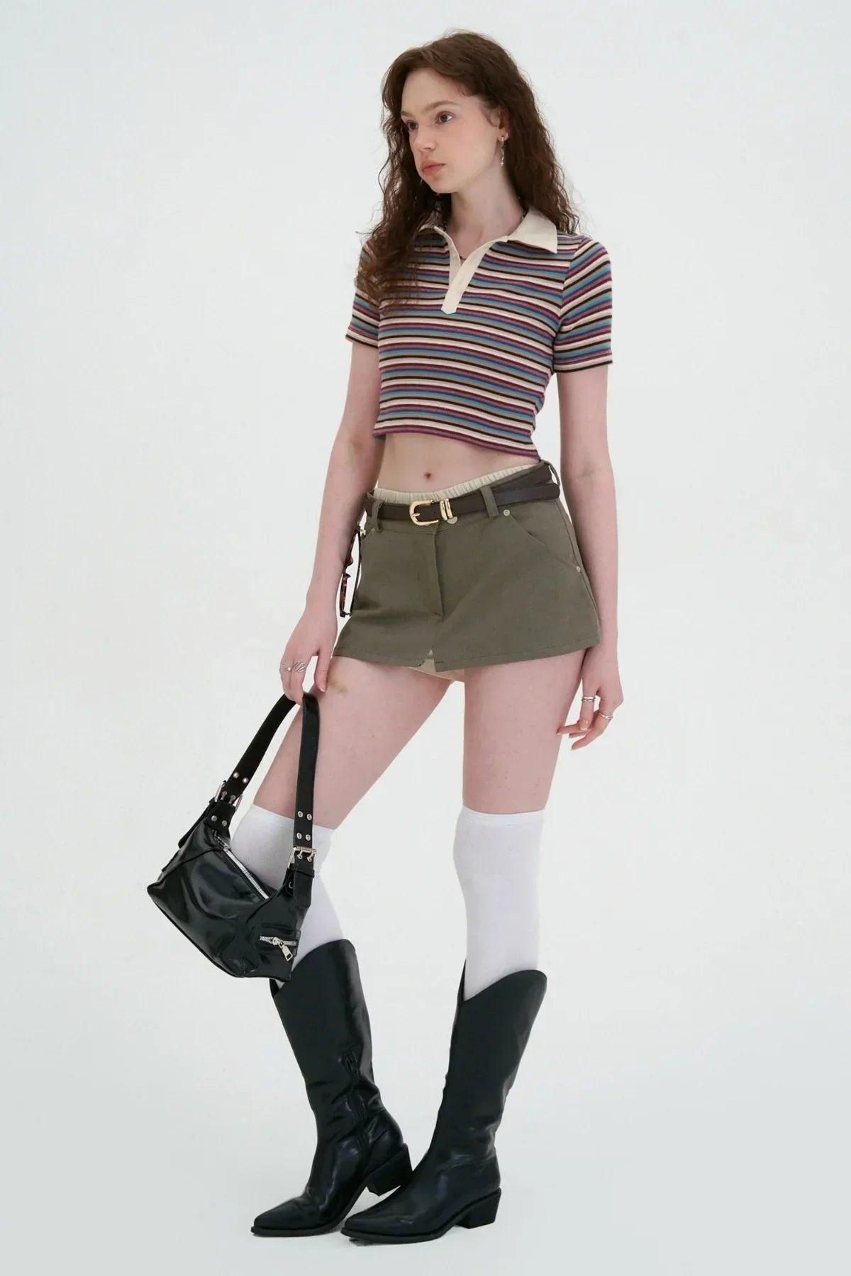 Woman wearing striped cropped polo shirt, olive green skort, white knee socks, black boots, and holding black handbag