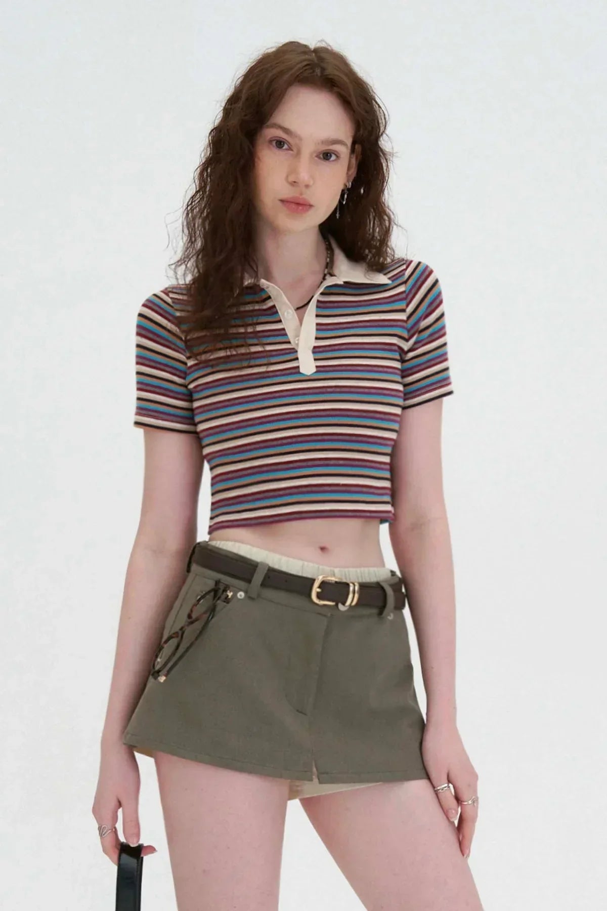 Young woman wearing striped cropped polo shirt and olive green skort with belt on white background