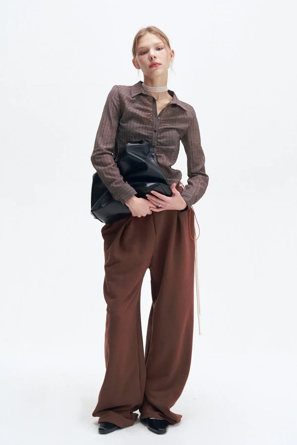 Model wearing brown striped button-up shirt and wide-leg brown pants, holding black bag on white background