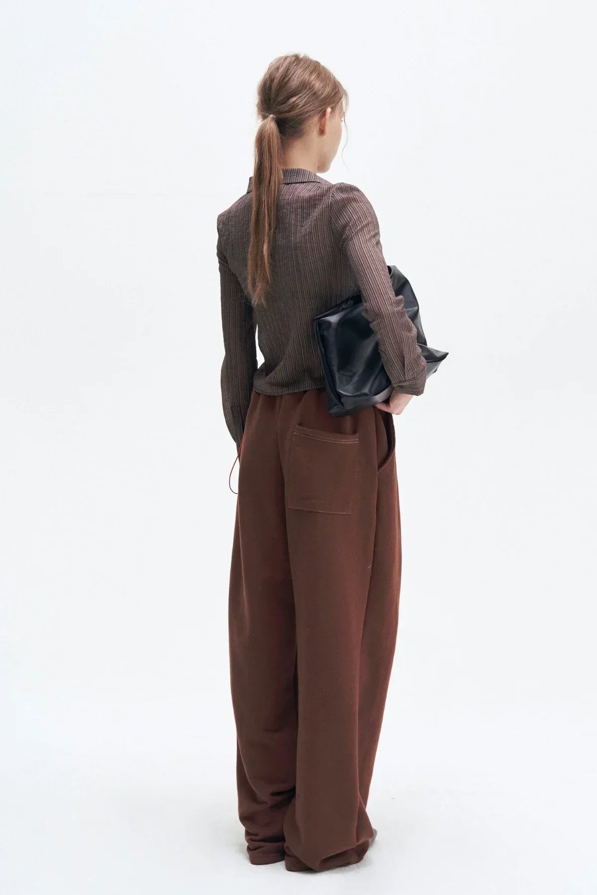 Woman wearing brown ribbed shirt and wide brown pants holding a black clutch bag on white background