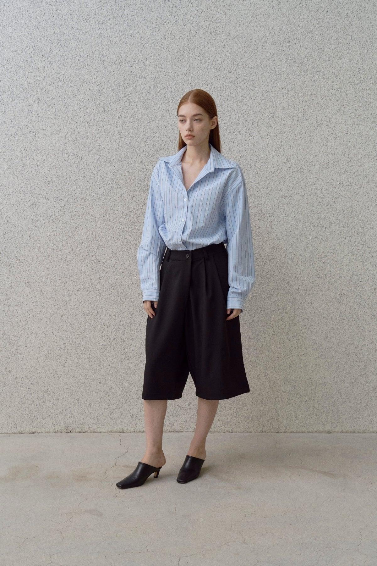 Woman wearing a blue striped button-up shirt, black wide-leg shorts, and black heeled mules against a textured wall