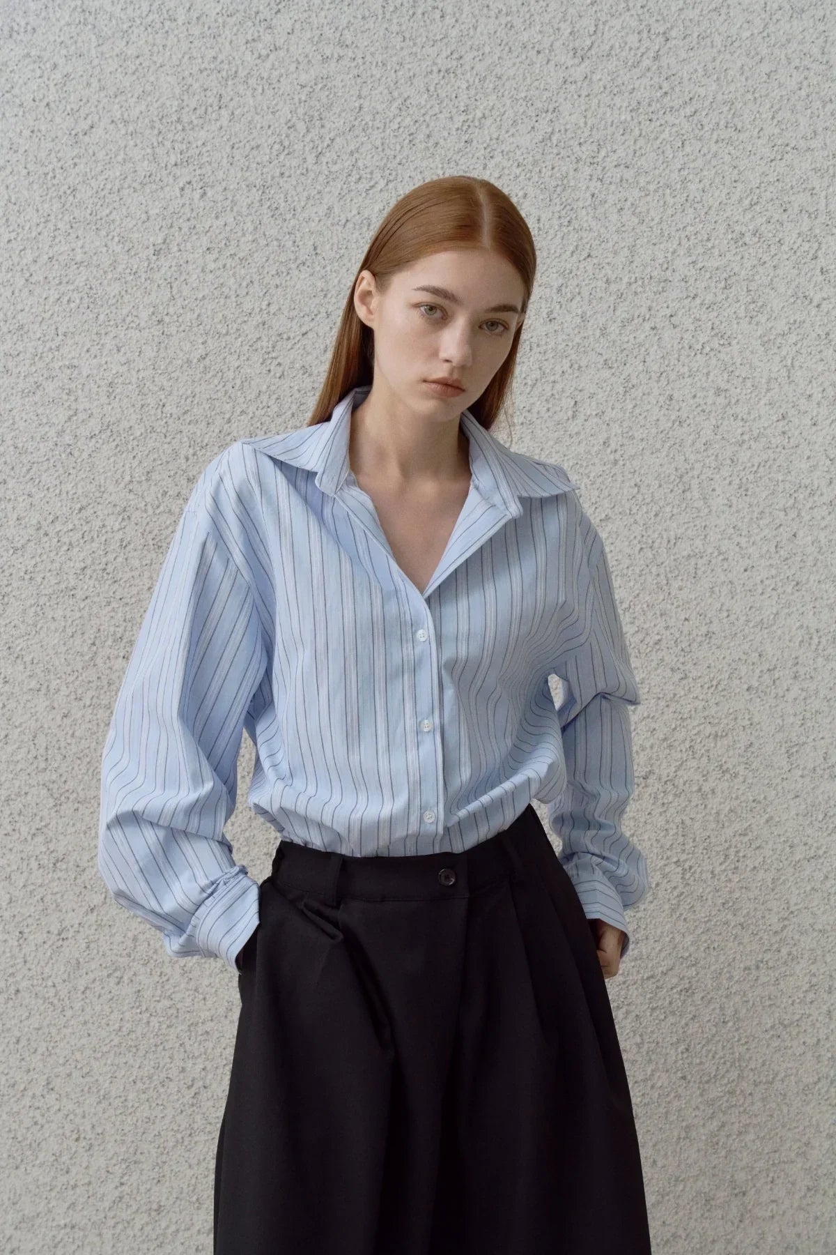 Woman wearing a blue striped button-up shirt and black high-waisted pants, standing against textured gray wall