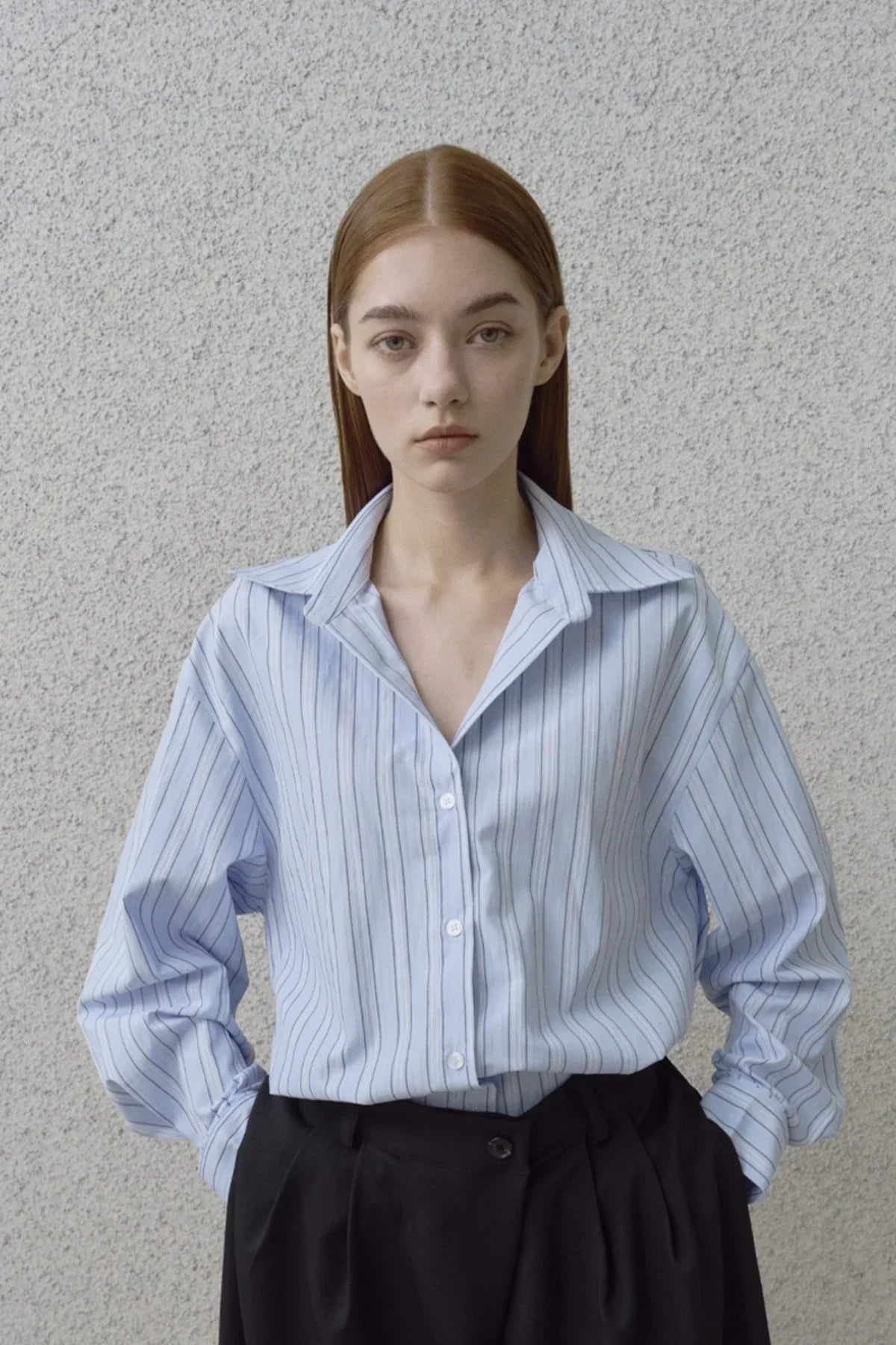 Young woman wearing light blue striped button-up shirt and black pants against textured gray wall