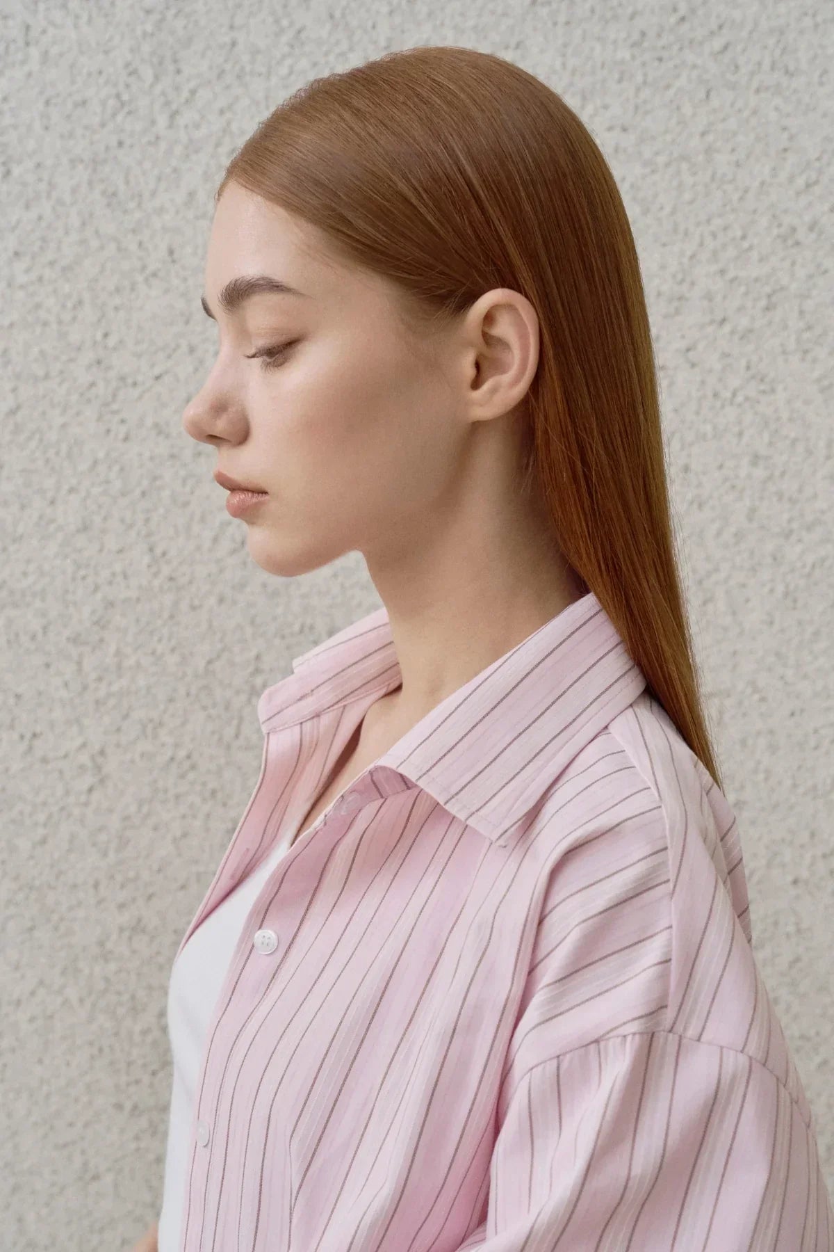 Profile of woman with straight red hair wearing pink striped shirt against textured wall