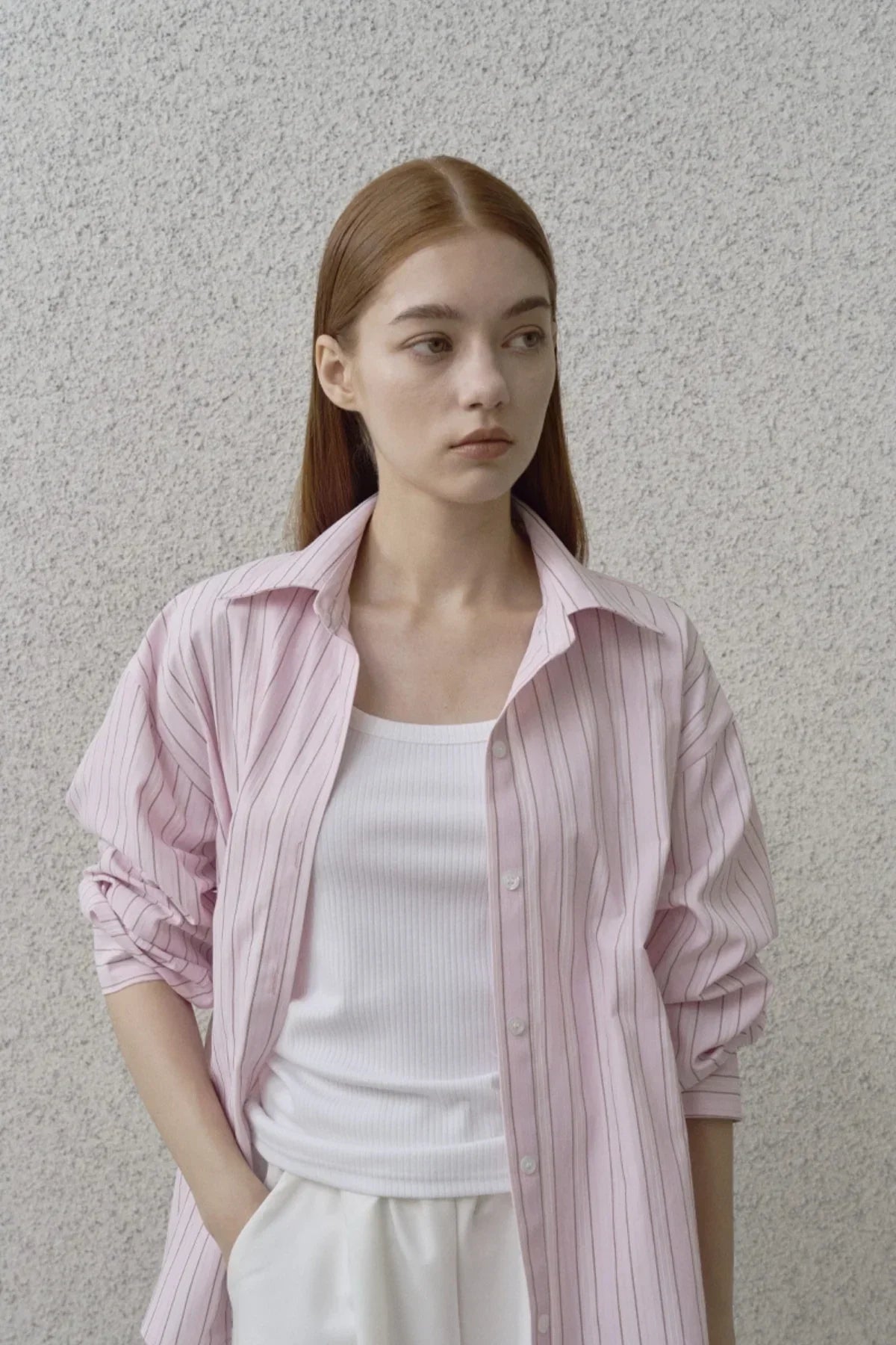 Young woman wearing pink striped shirt over white tank top and white pants against textured wall