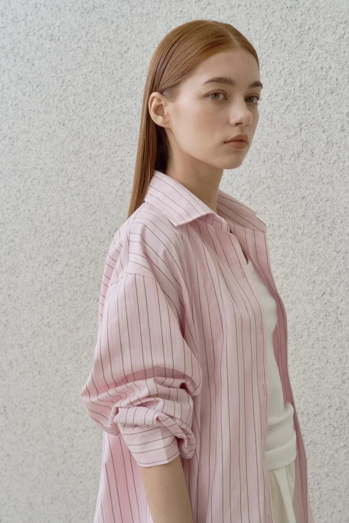 Young woman with straight red hair wearing a loose pink striped shirt over a white top against a light textured wall