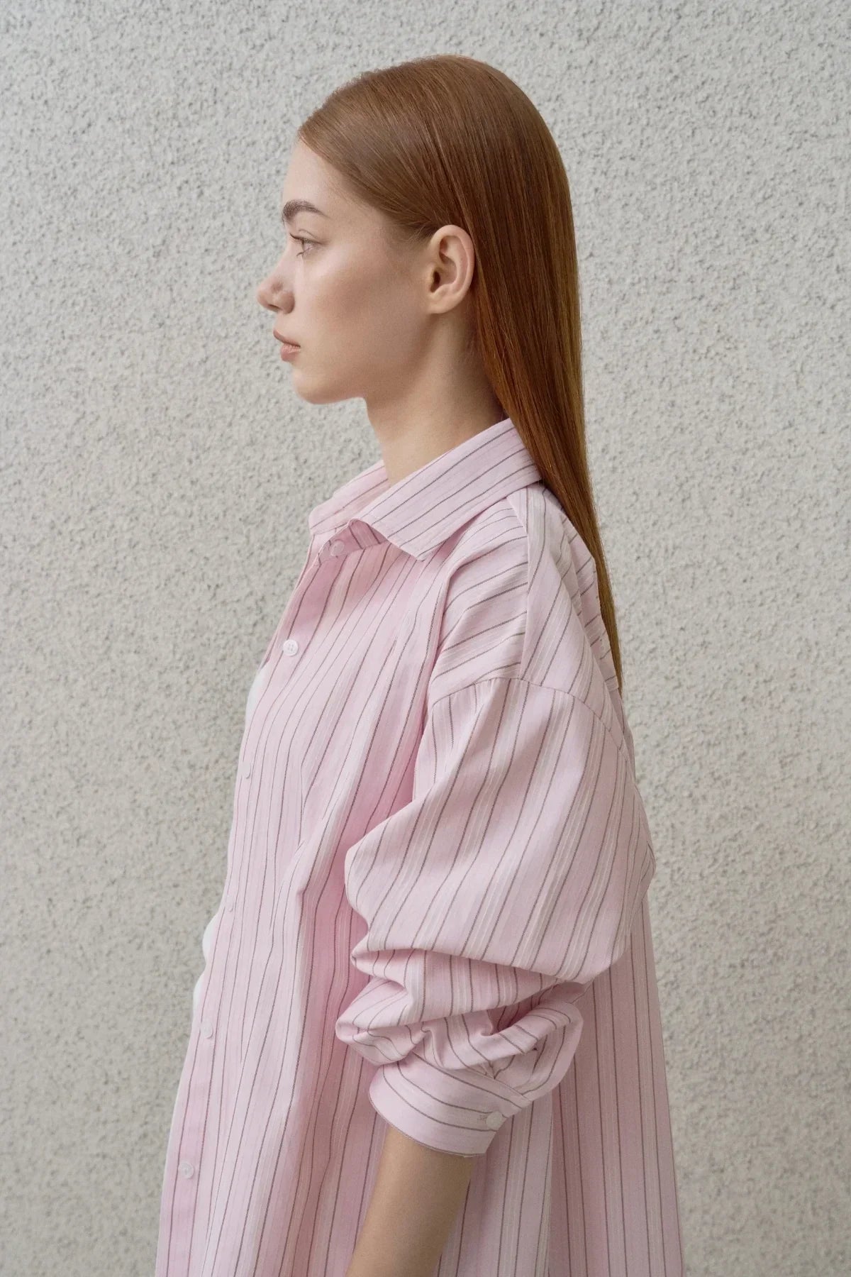Profile of woman with straight red hair wearing a pink striped button-up shirt against a textured wall