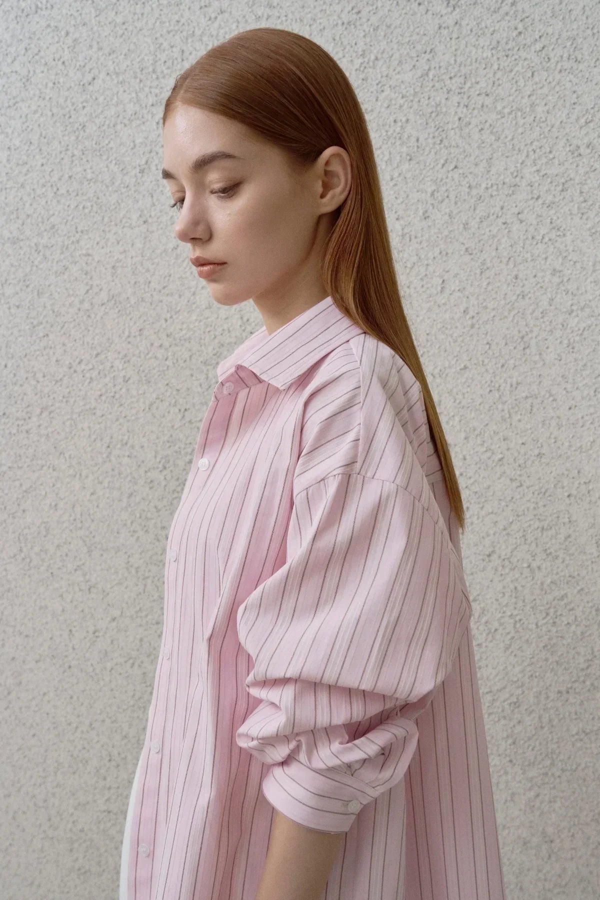 Profile of young woman with straight red hair wearing a loose pink striped button-up shirt against textured gray wall