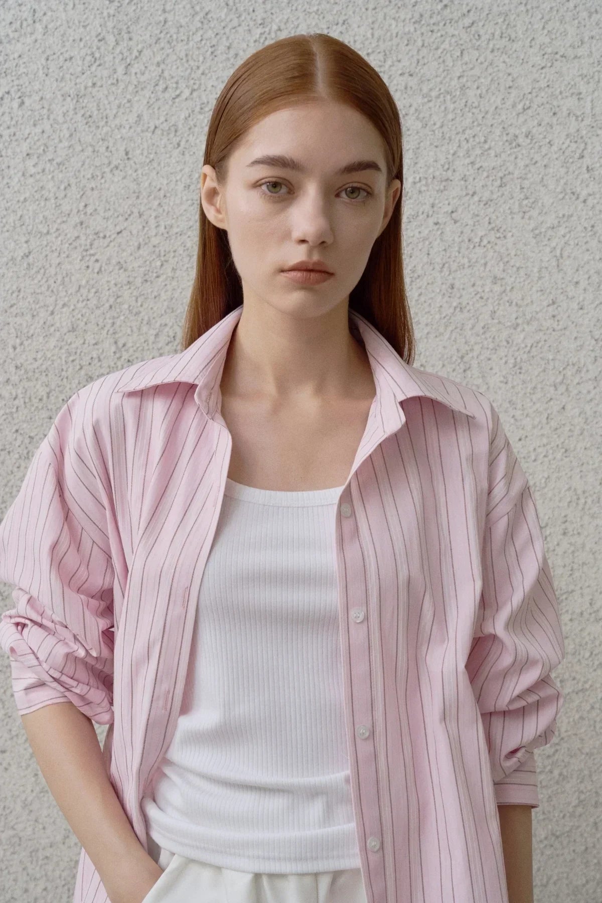 Young woman wearing pink striped shirt over white ribbed tank top against textured wall