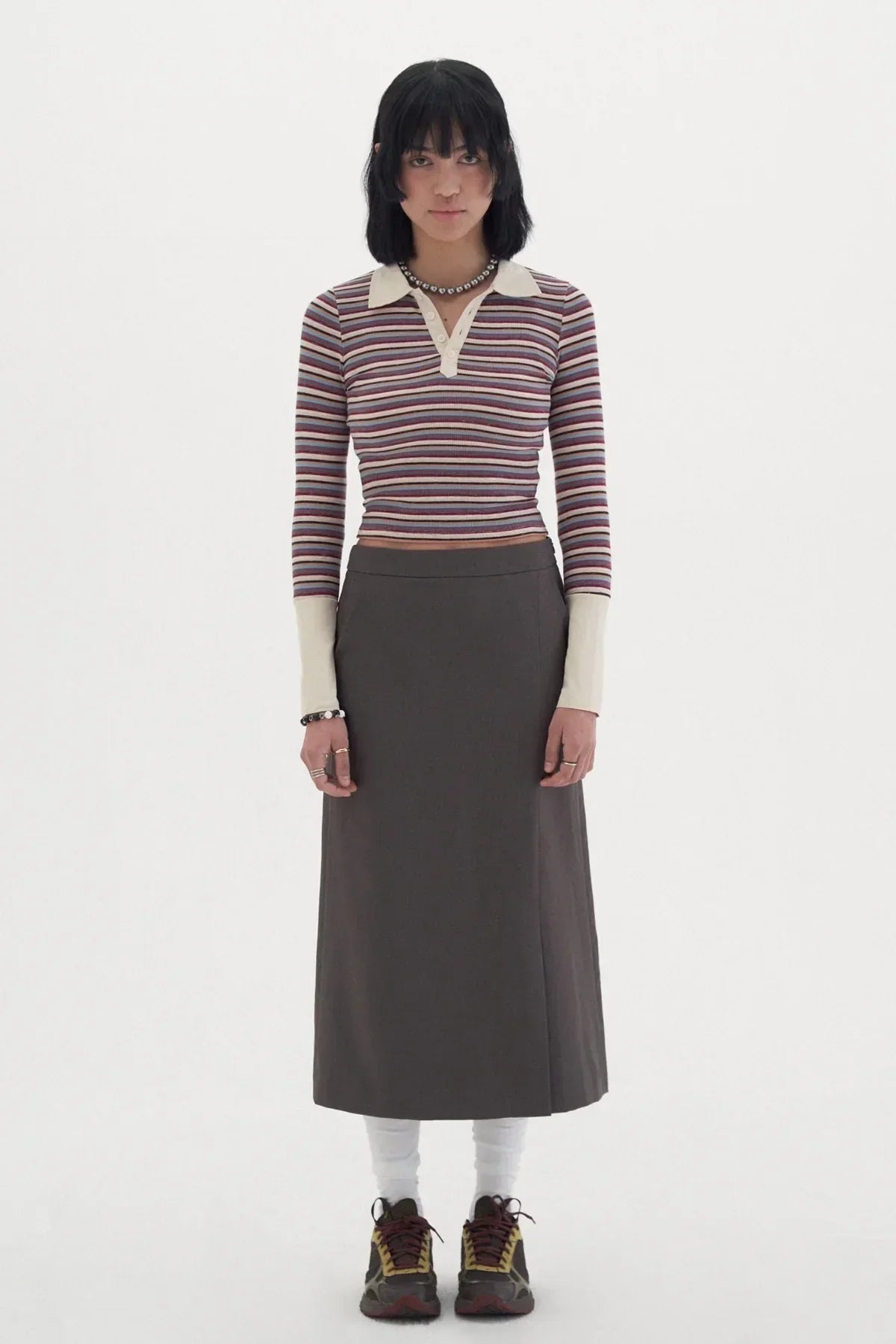 Woman wearing striped long-sleeve polo shirt, brown midi skirt, white socks, and sneakers on white background