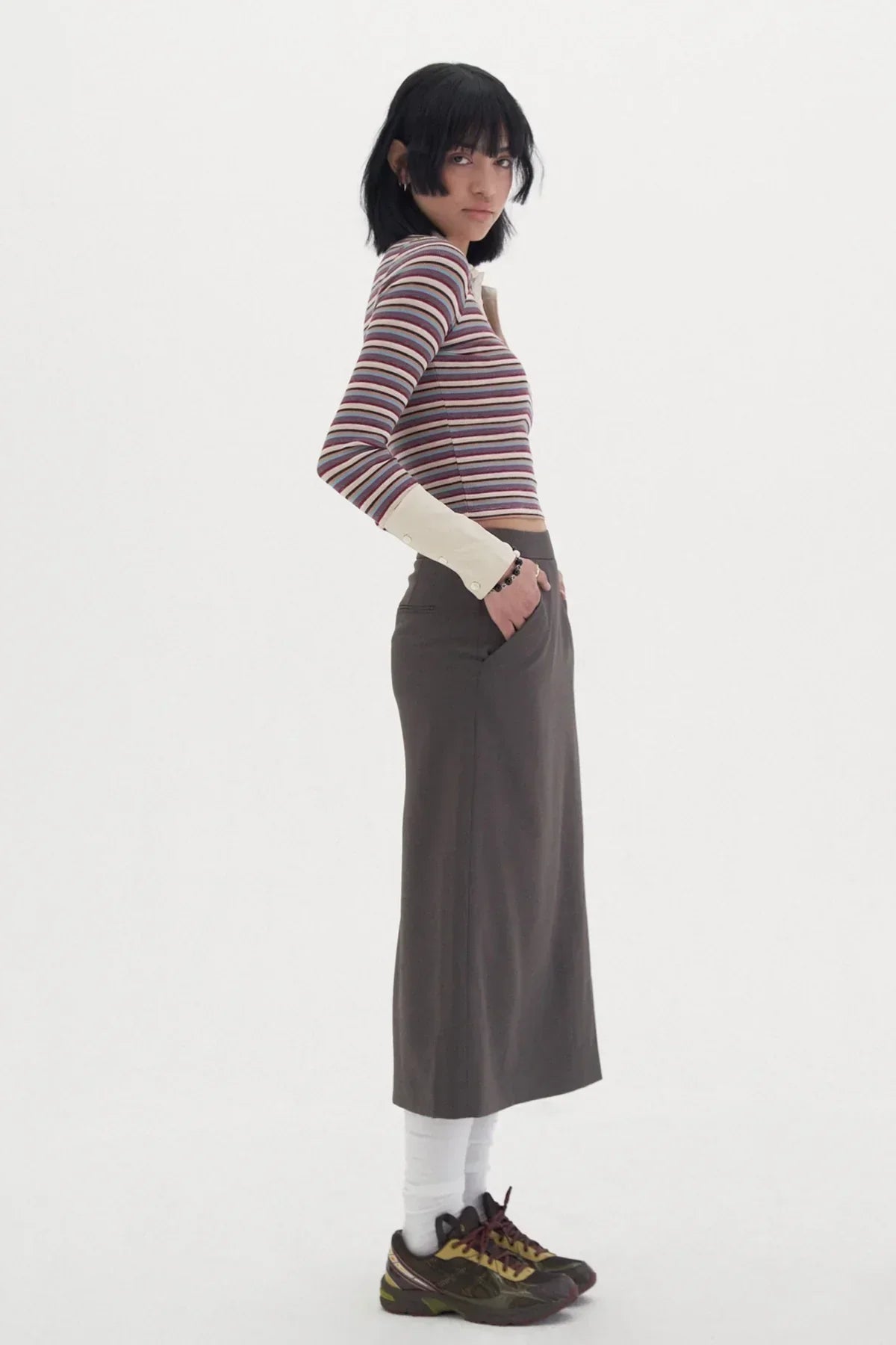 Side view of a woman wearing a striped long-sleeve top, dark midi skirt, white socks, and black sneakers