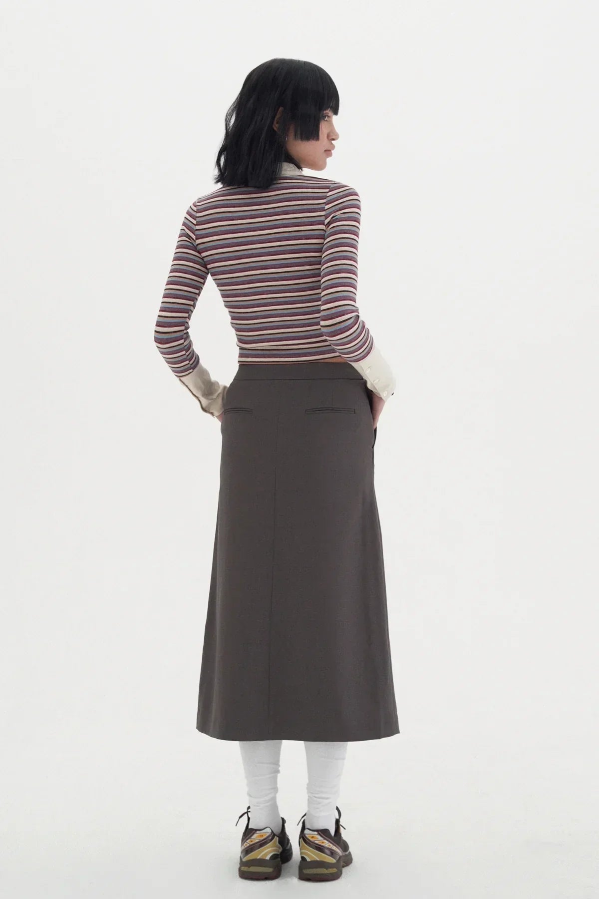 Woman in a striped long-sleeve polo, gray midi skirt, white leggings, and sneakers on white background