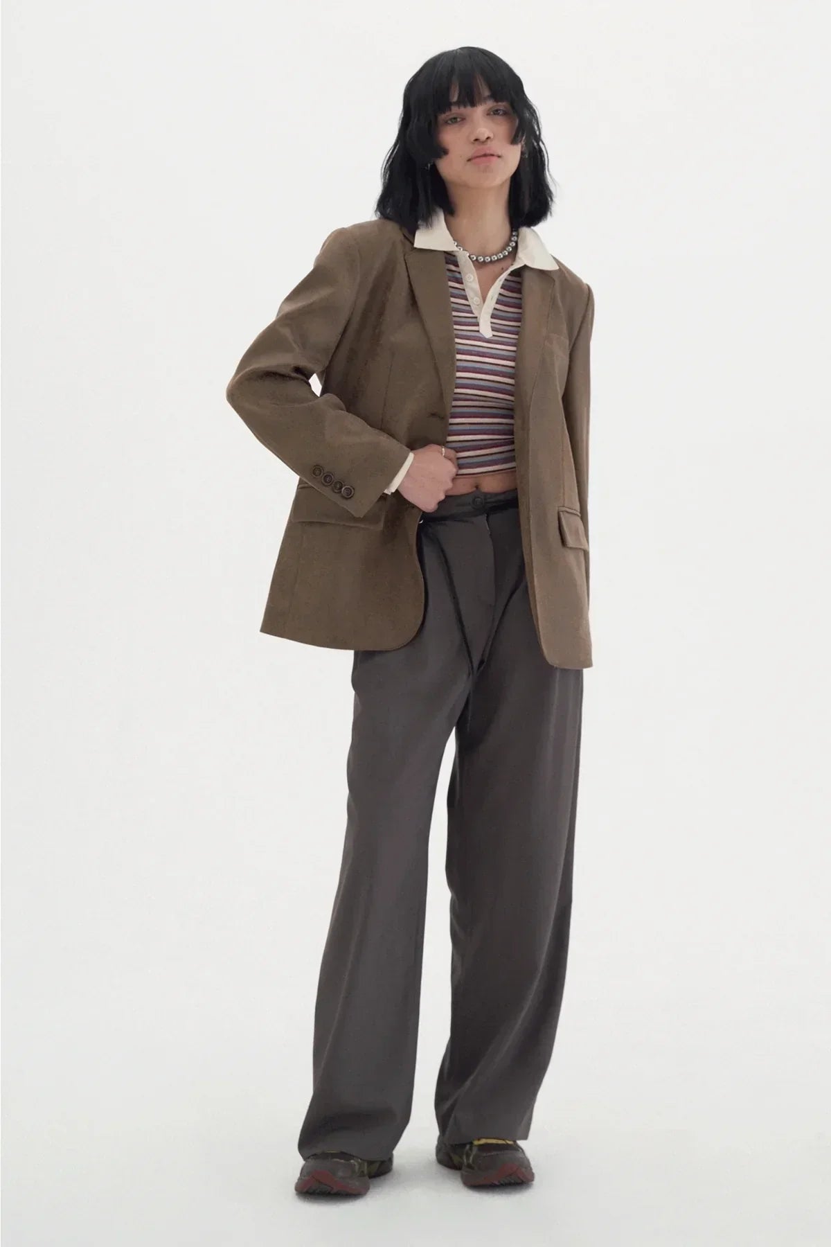 Woman wearing striped polo shirt, brown blazer, wide-leg gray pants, and sneakers on white background