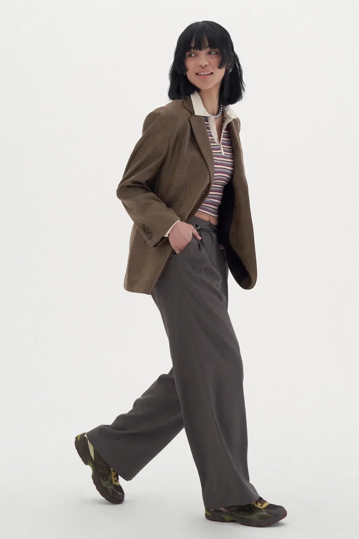 Woman wearing striped polo shirt, brown blazer, wide-leg gray pants, and sneakers walking in studio