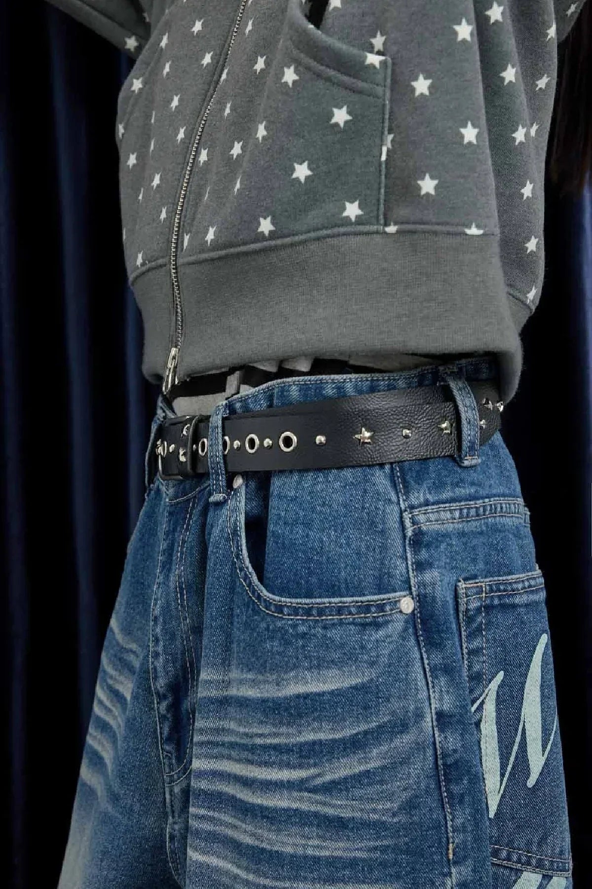 Close-up of person wearing star-patterned grey hoodie, studded black leather belt, and blue jeans with light distressed washes.