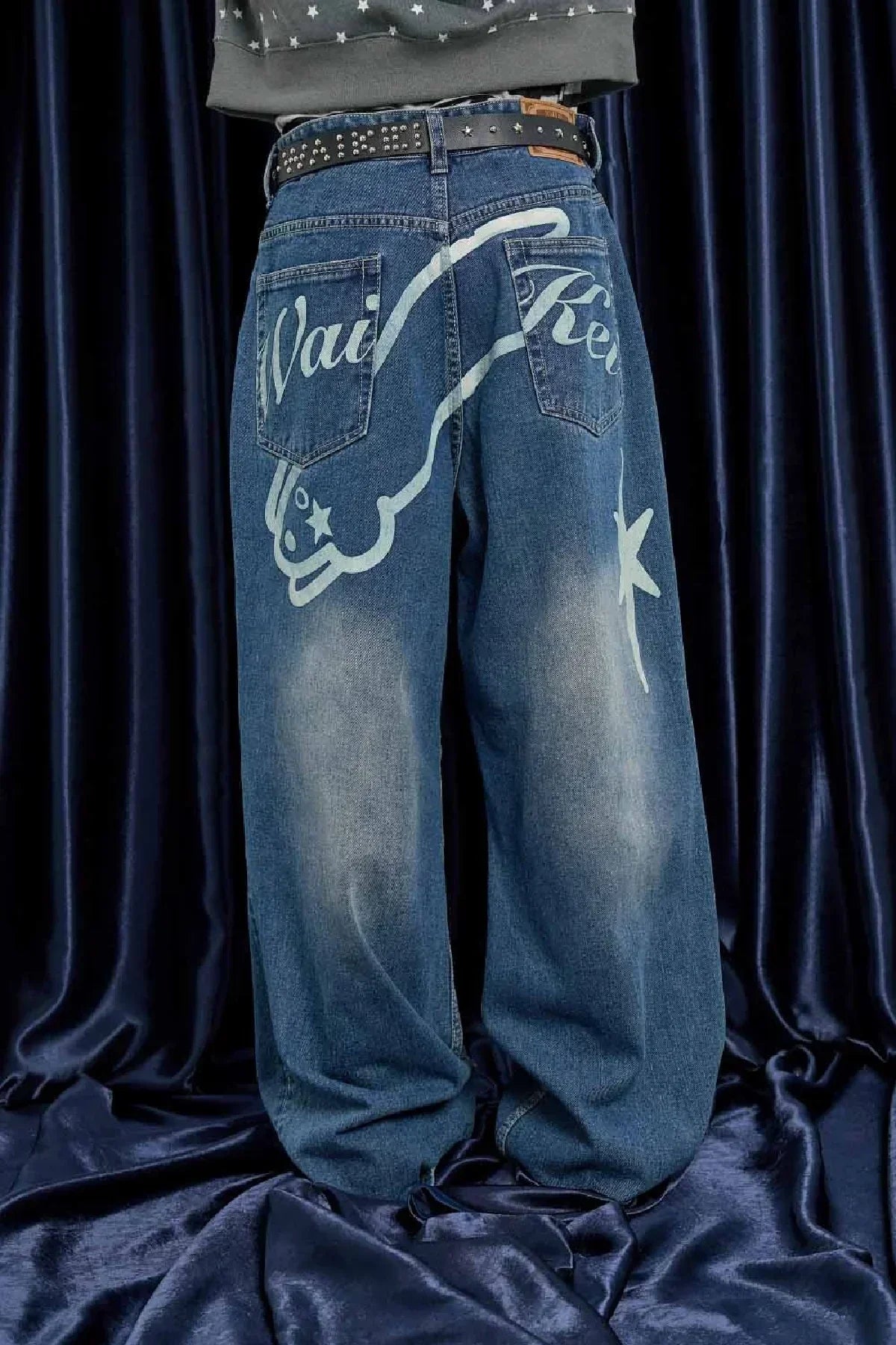 Back view of baggy blue jeans with white graphic and studded black belt on dark fabric background