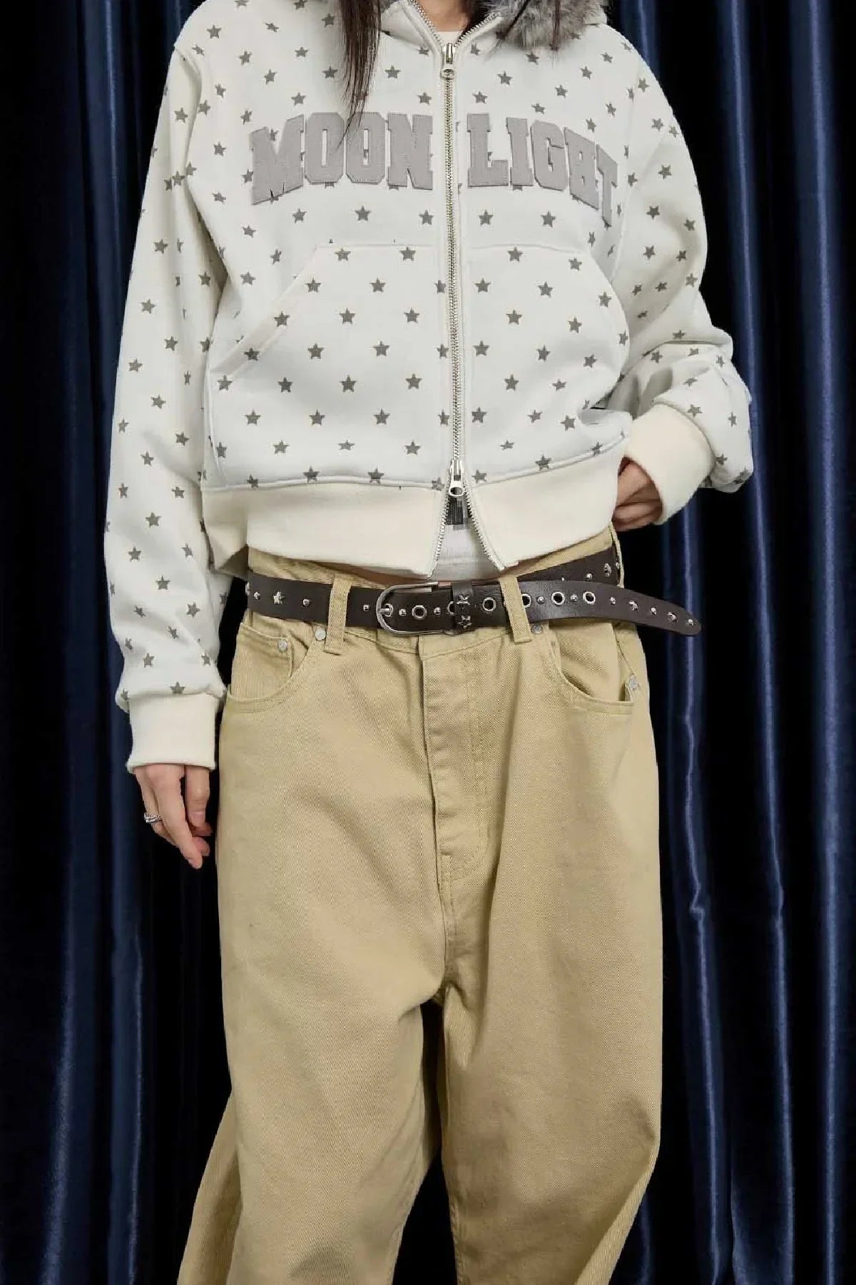 Person wearing beige pants with a brown studded leather belt and a white zip-up hoodie with gray star patterns and 'MOON LIGHT' text