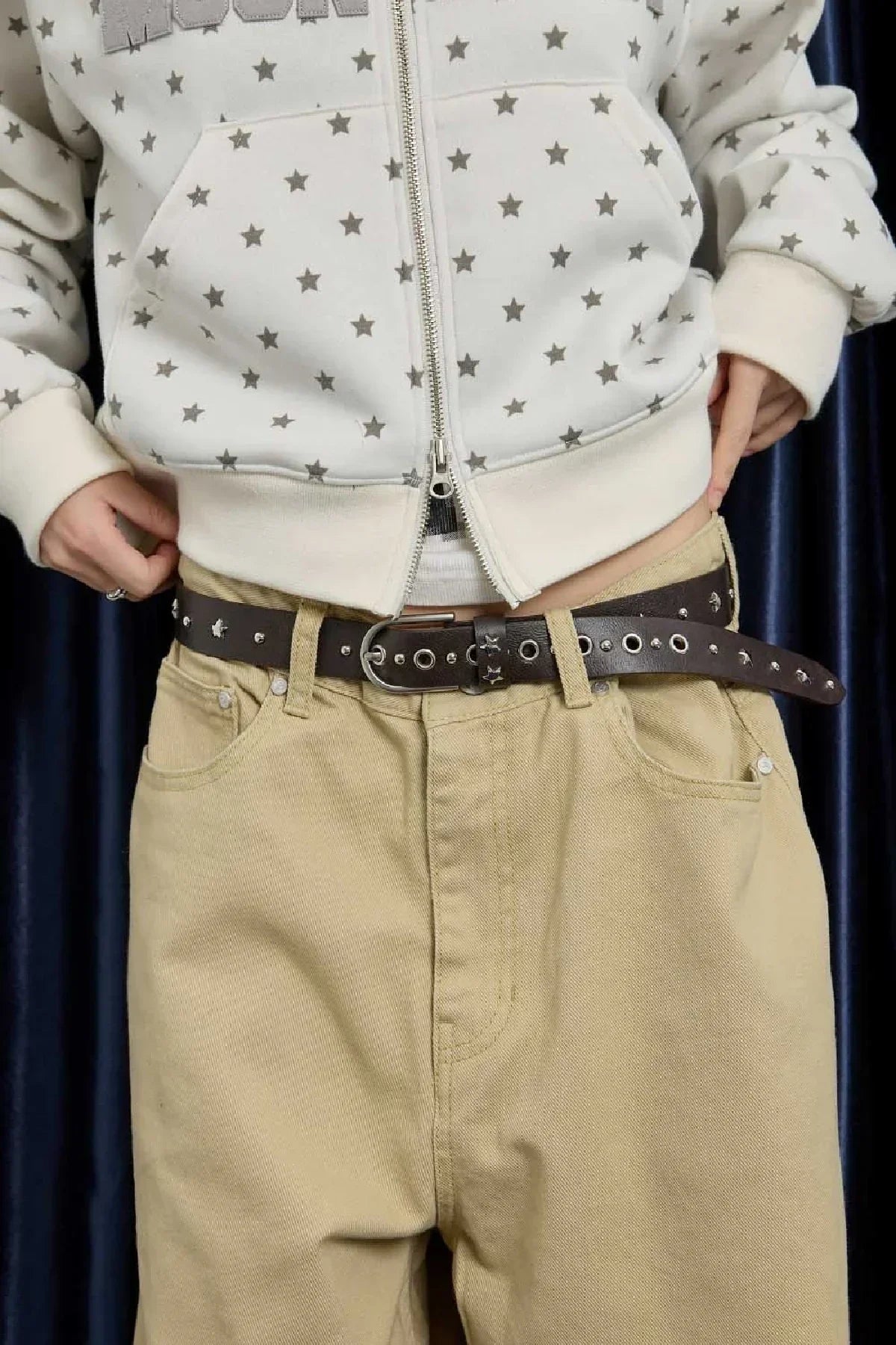 Person wearing beige pants with a brown studded belt and white star-patterned zip-up hoodie