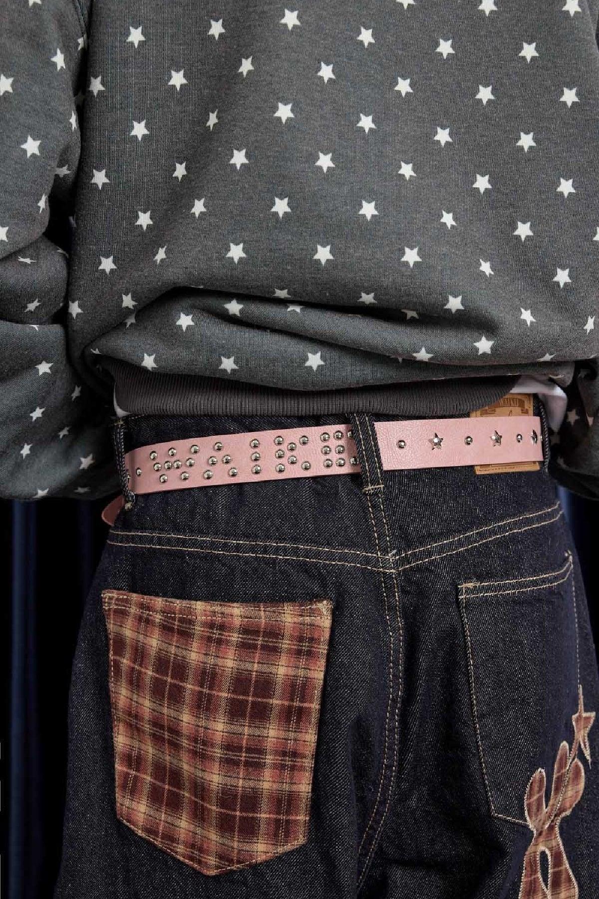 Close-up of person wearing dark denim jeans with plaid patch pocket and pink studded belt featuring star details