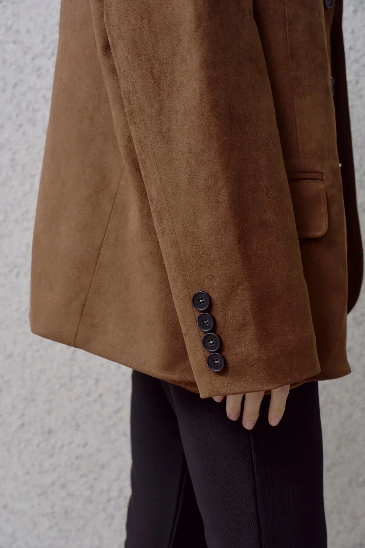 Close-up of brown suede blazer sleeve with four black buttons, worn with black pants
