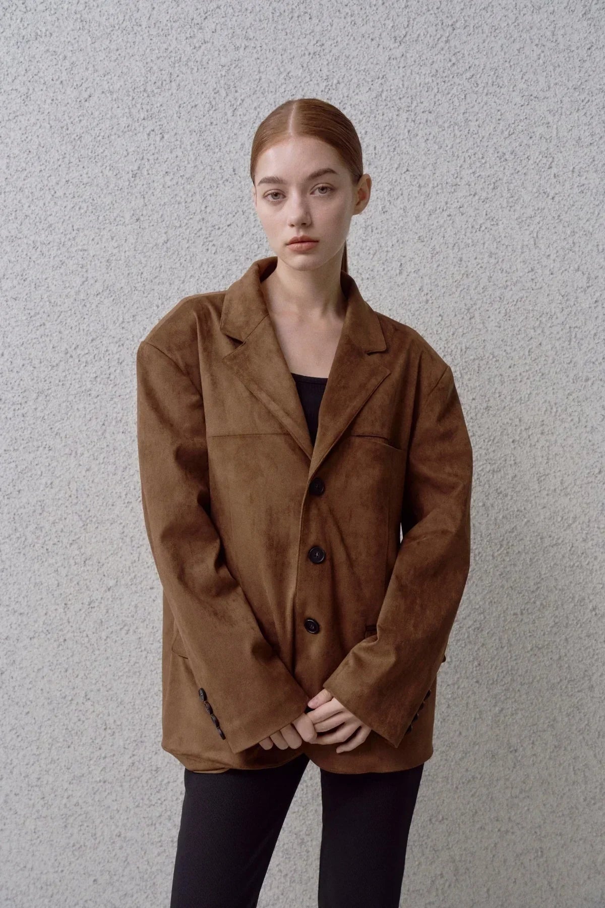 Young woman wearing a brown suede oversized blazer with black pants against a gray textured wall