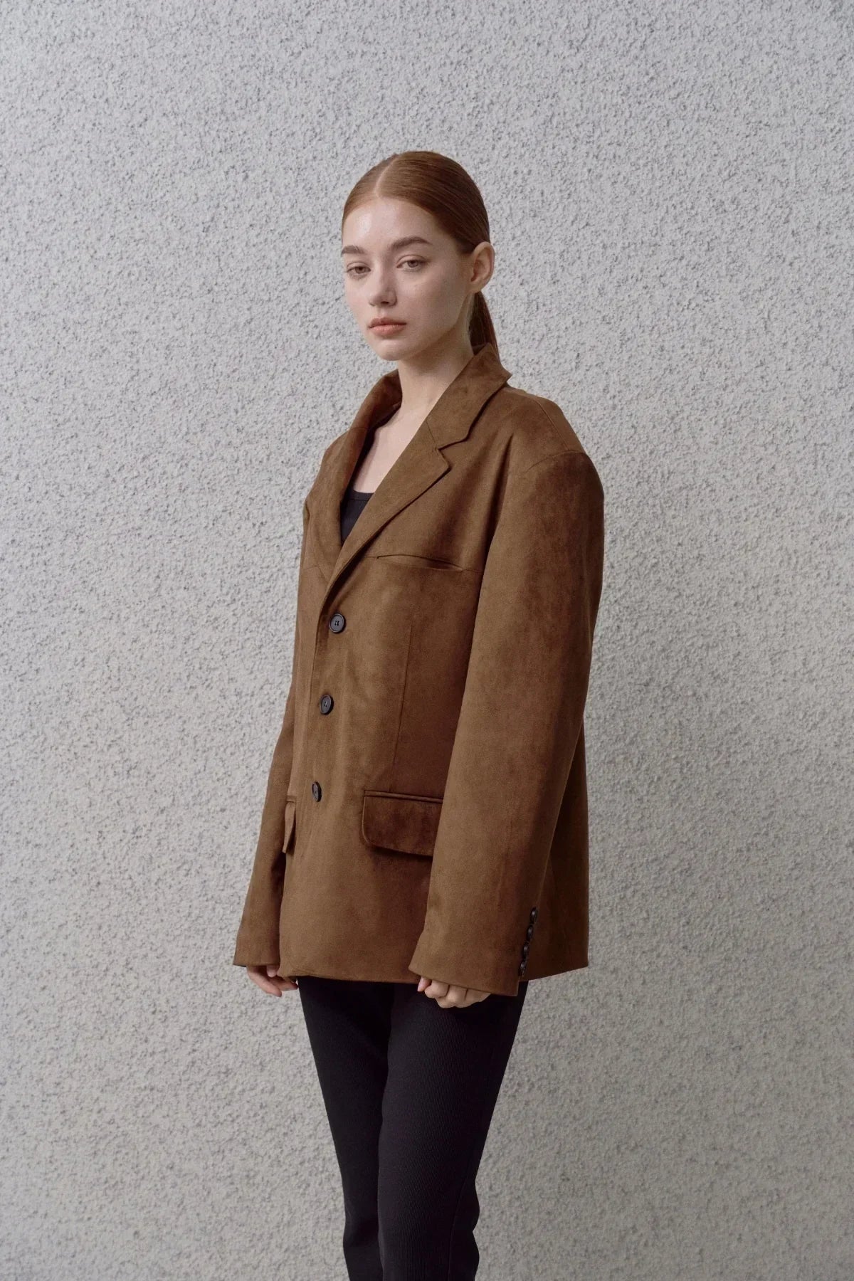 Young woman wearing a brown suede blazer with black pants against gray textured wall
