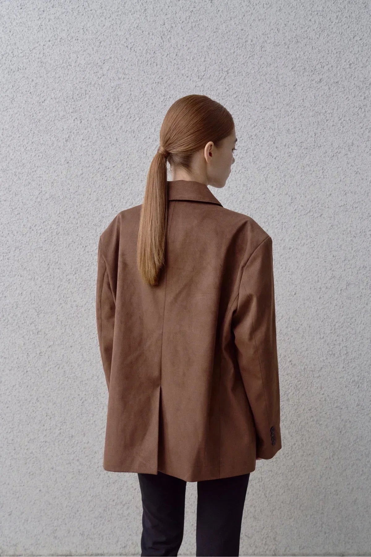 Woman with long ponytail wearing brown suede blazer standing against textured gray wall