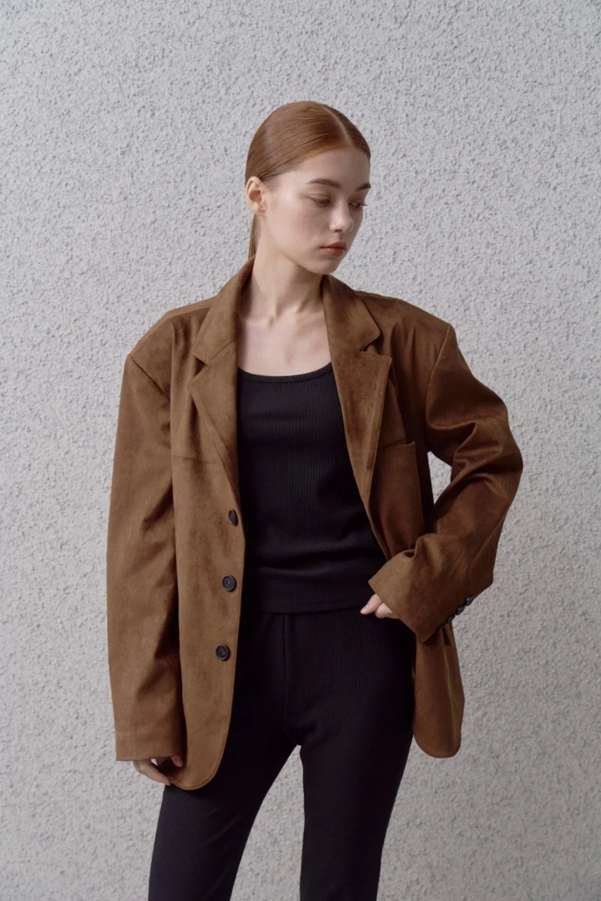 Young woman wearing a brown suede blazer over a black tank top and black pants standing against a textured gray wall