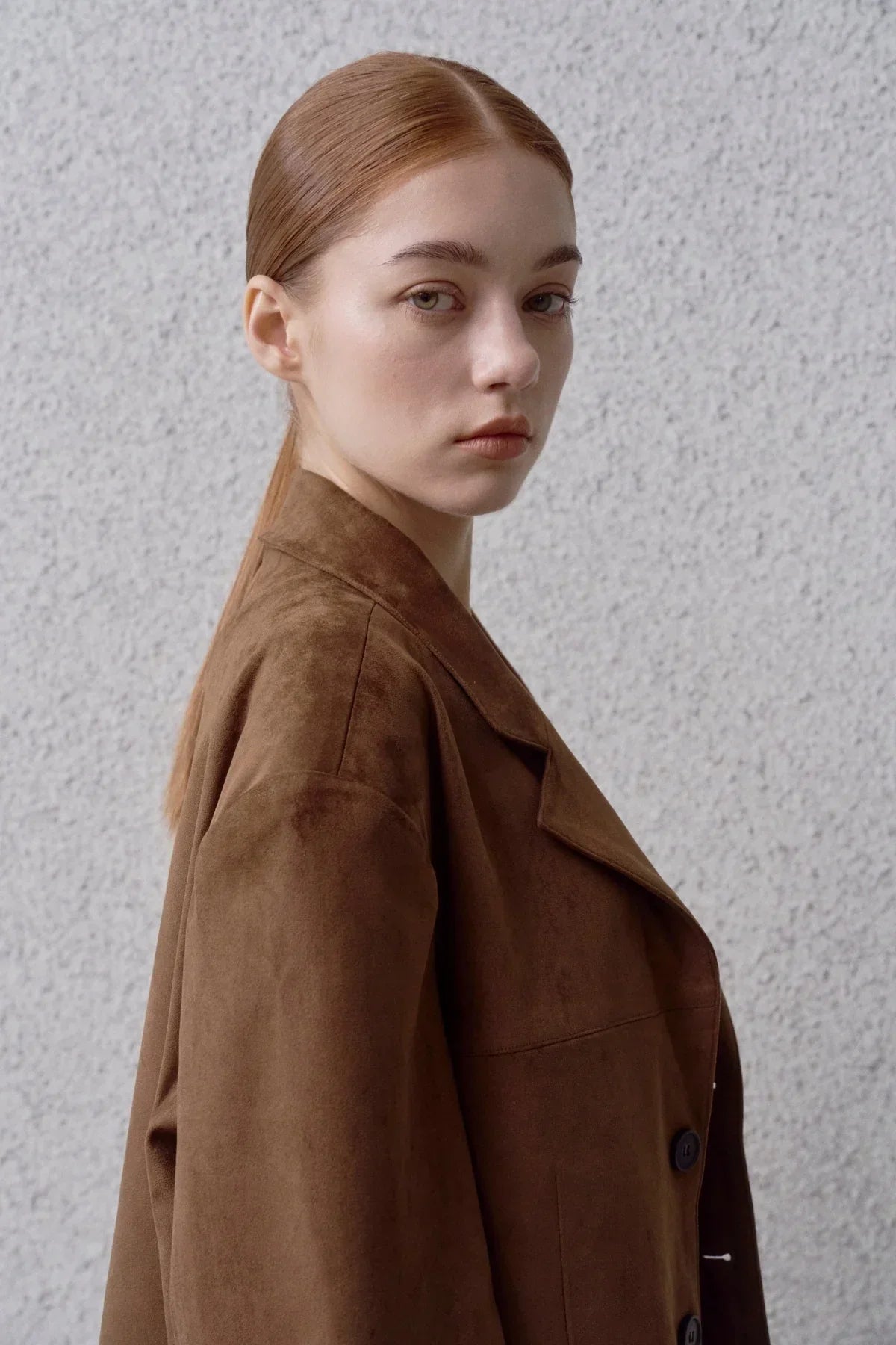 Young woman with red hair wearing a brown suede blazer against a textured gray wall