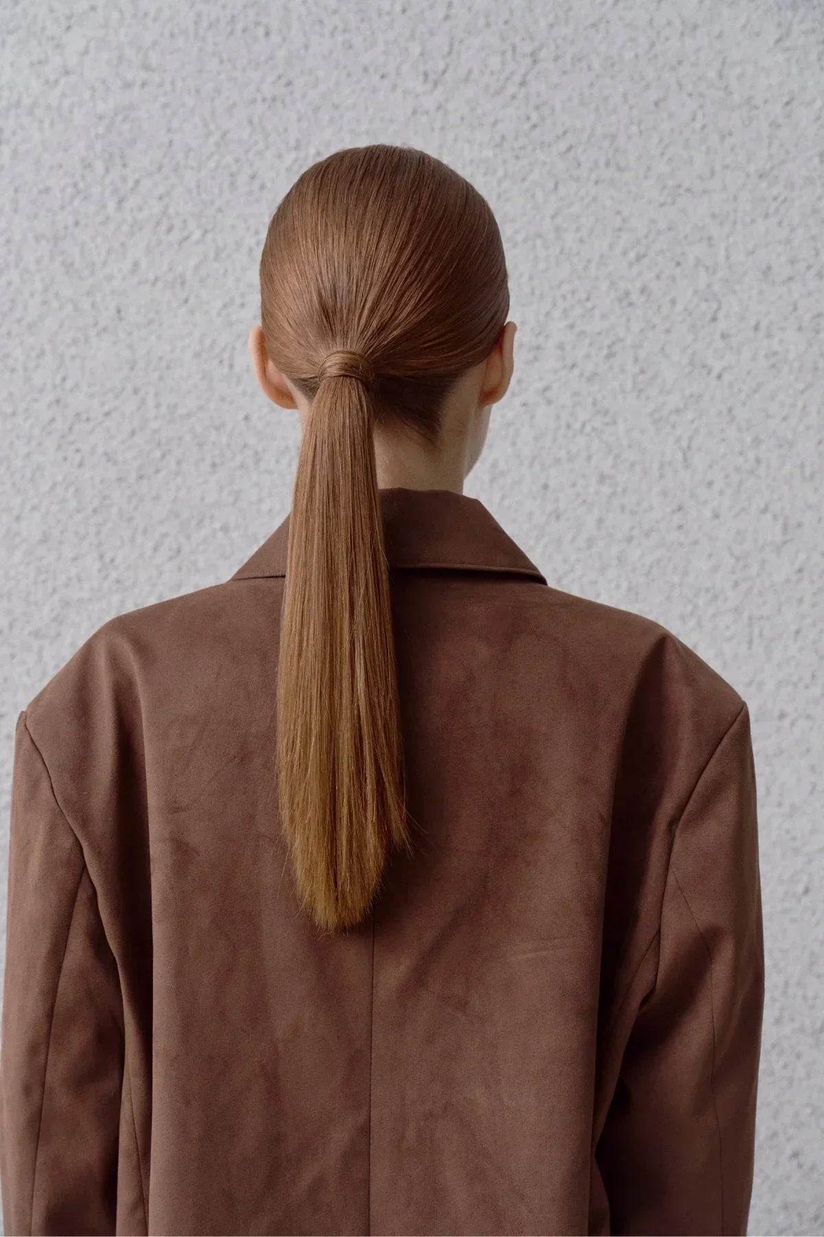 Back view of a woman with sleek brown ponytail wearing a brown suede blazer against a light textured wall