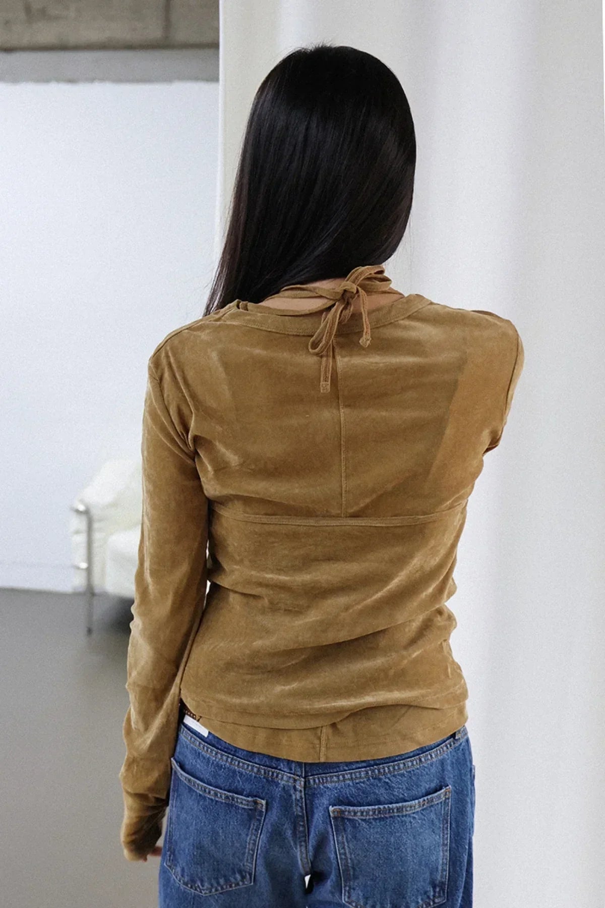 Woman with long dark hair wearing camel suede long-sleeve top and blue jeans, back view indoors