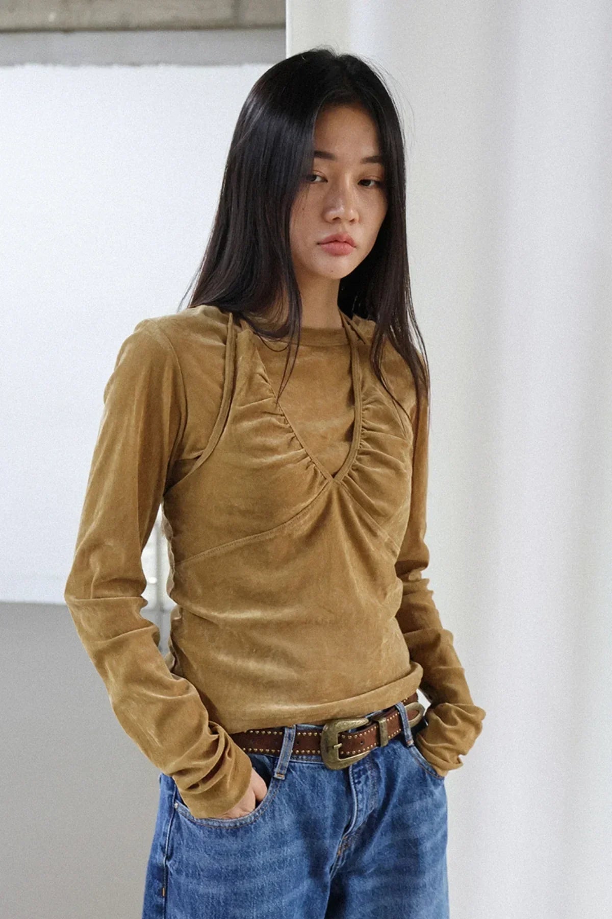 Woman wearing camel suede long sleeve top with ruched front and blue jeans standing indoors
