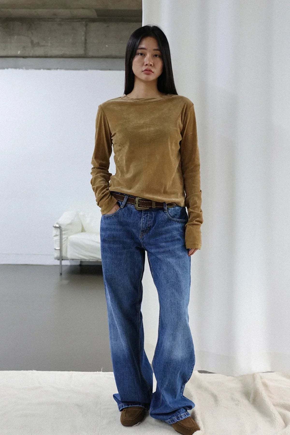 Woman wearing camel suede long sleeve top, blue jeans, and brown loafers in minimal indoor setting
