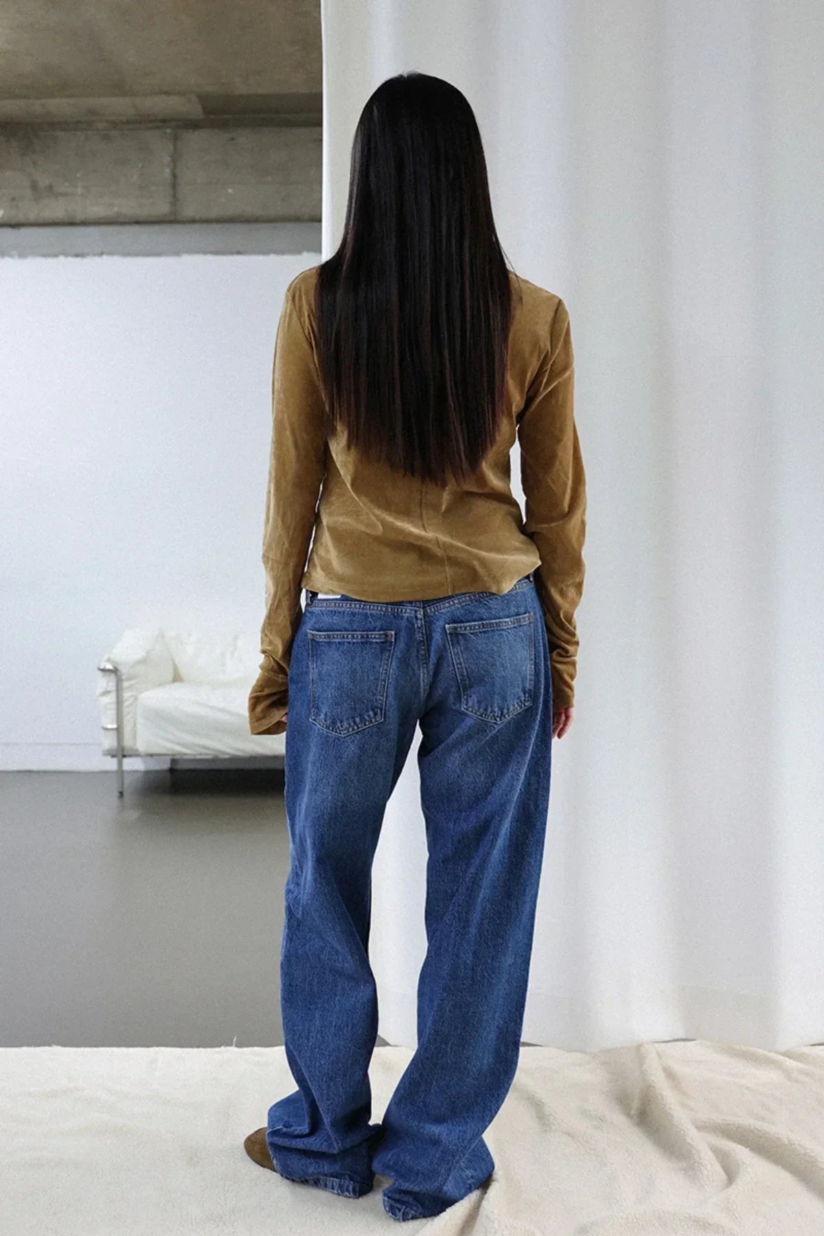 Woman with long straight hair wearing camel suede long sleeve top and loose blue jeans standing indoors