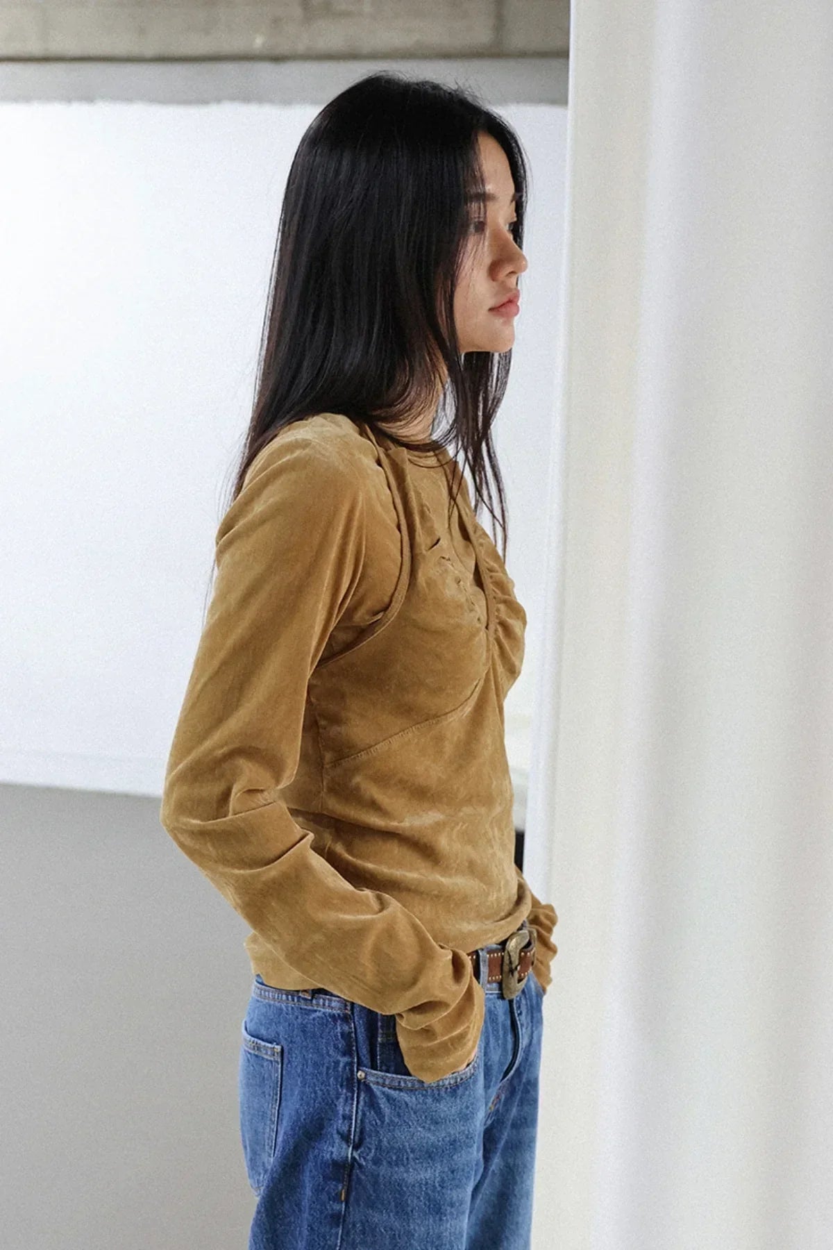Woman in camel suede long-sleeve top and blue jeans standing indoors against white wall