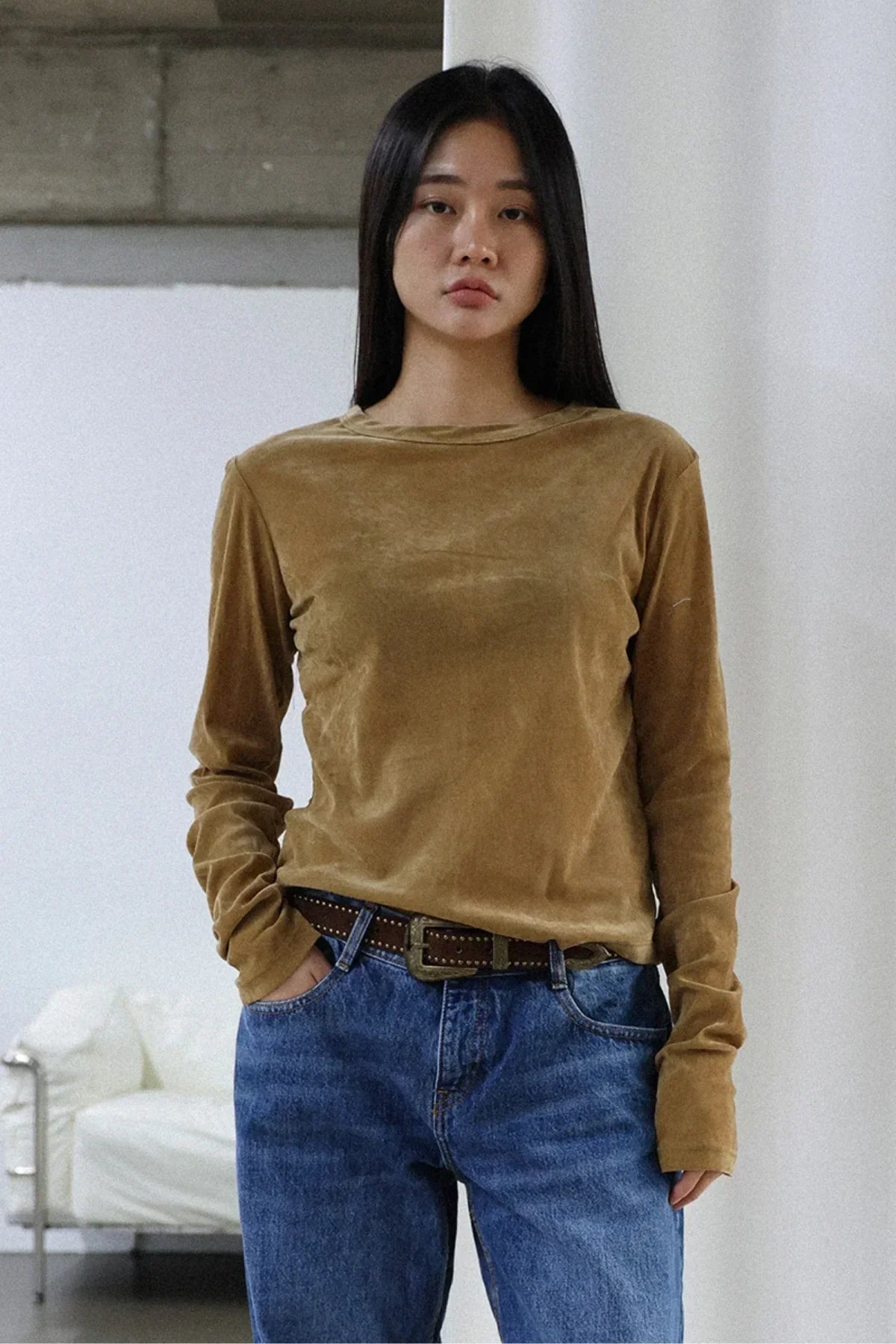 Woman wearing camel suede long sleeve top and blue jeans with brown belt in minimal indoor studio