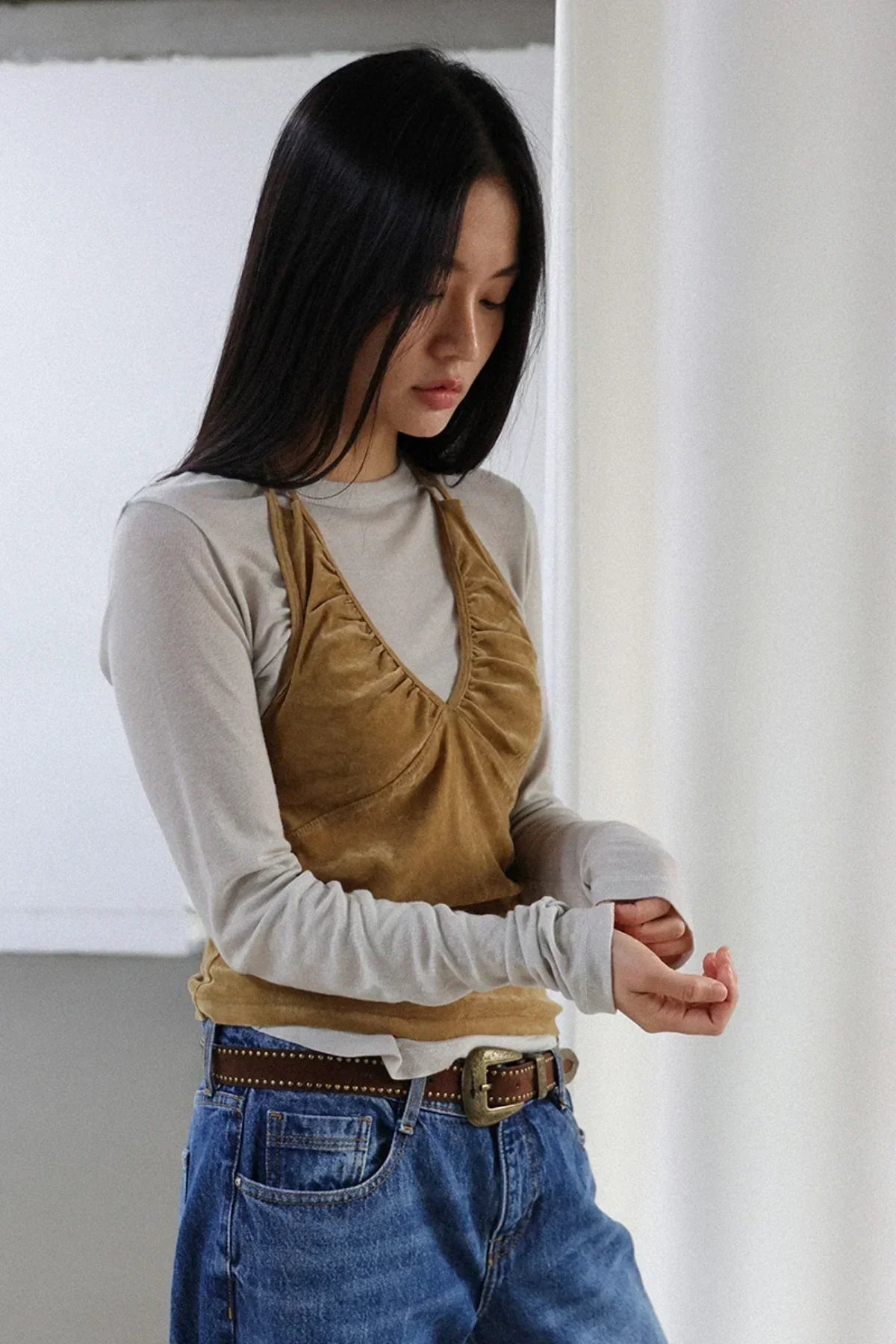 Woman wearing camel suede shirring bra top over light gray long sleeve shirt with blue jeans and brown belt